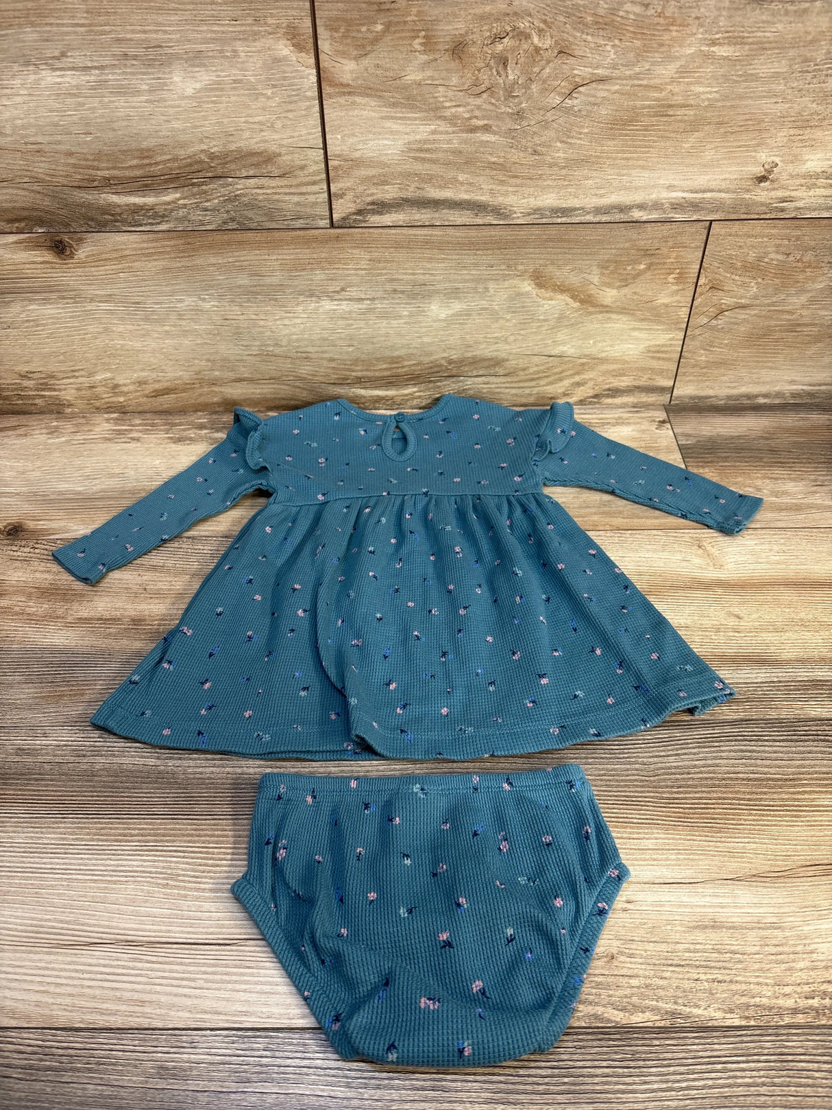 Flat lay of teal waffle-knit baby dress and bloomers, small pink floral print and ruffle shoulders on wood