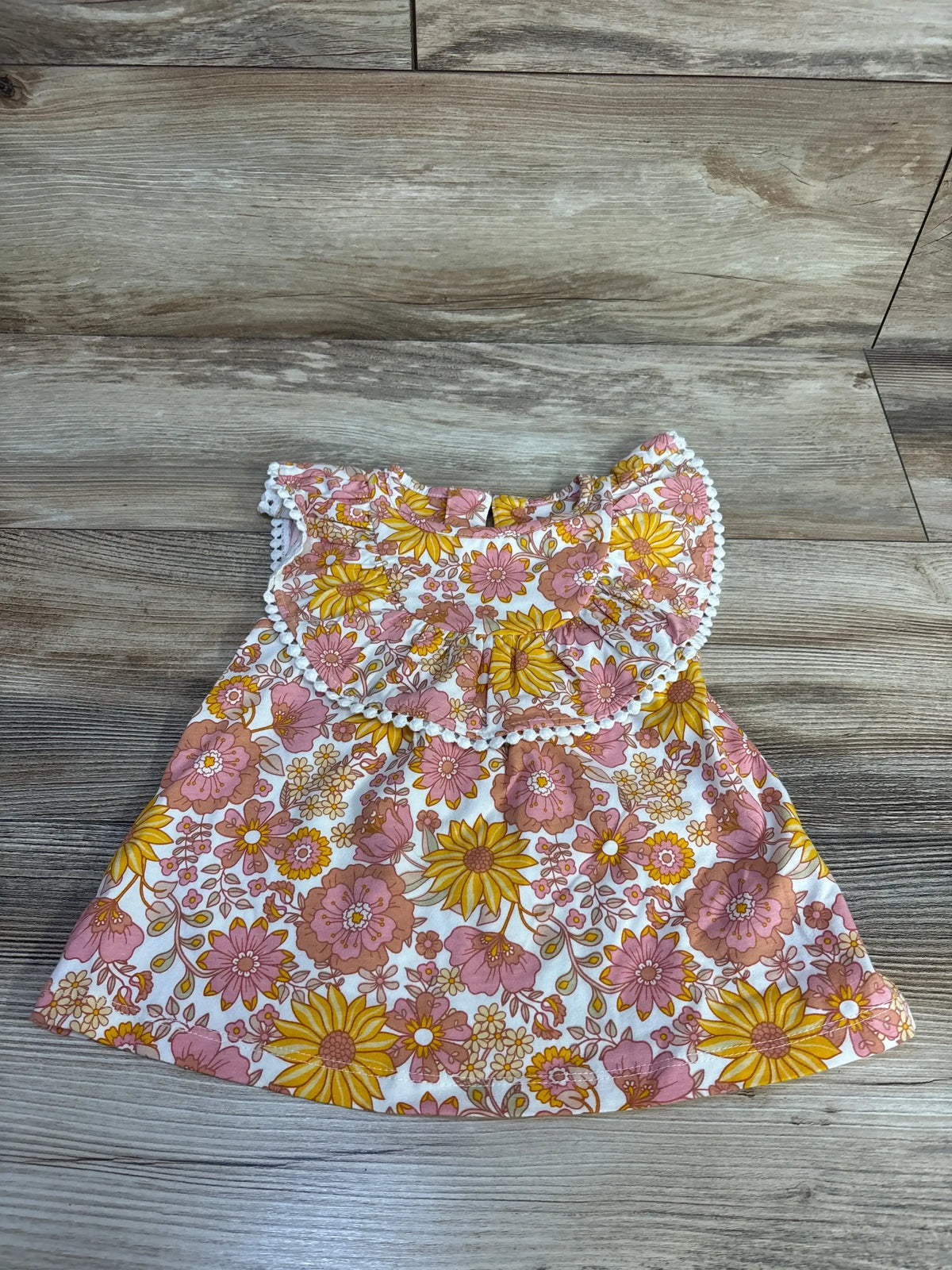 Flat lay of pink floral baby shirt, size 12 months, showing ruffled bib collar with pom-pom trim on light wood background