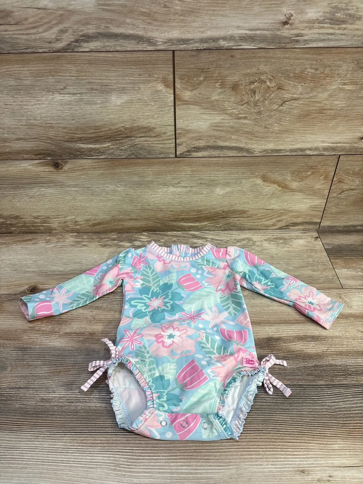 Front view of Ruffle Butts In Bloom rashguard swimsuit with long sleeves, blue floral print, ruffled legs and snaps on wood