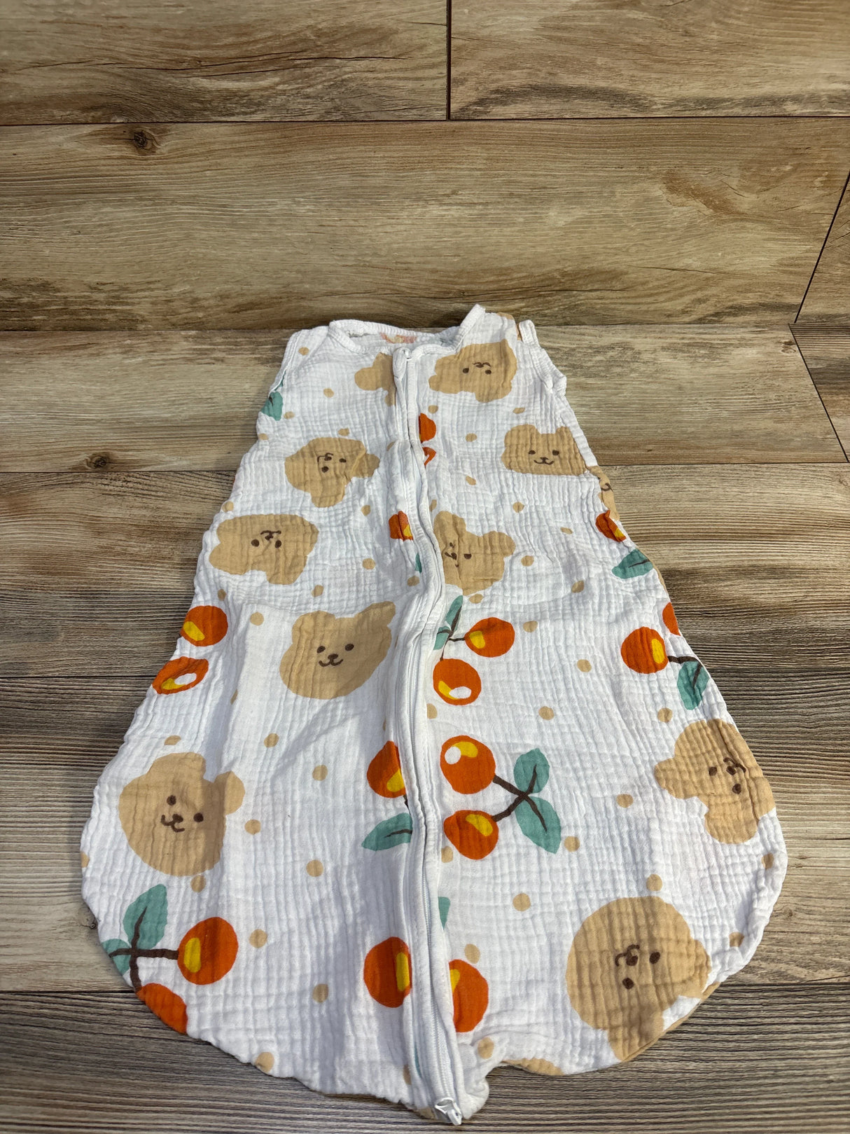 Top-down view of white gauze baby sleep sack (small 0-6m) with front zipper, bear face and orange fruit print laid on wood surface