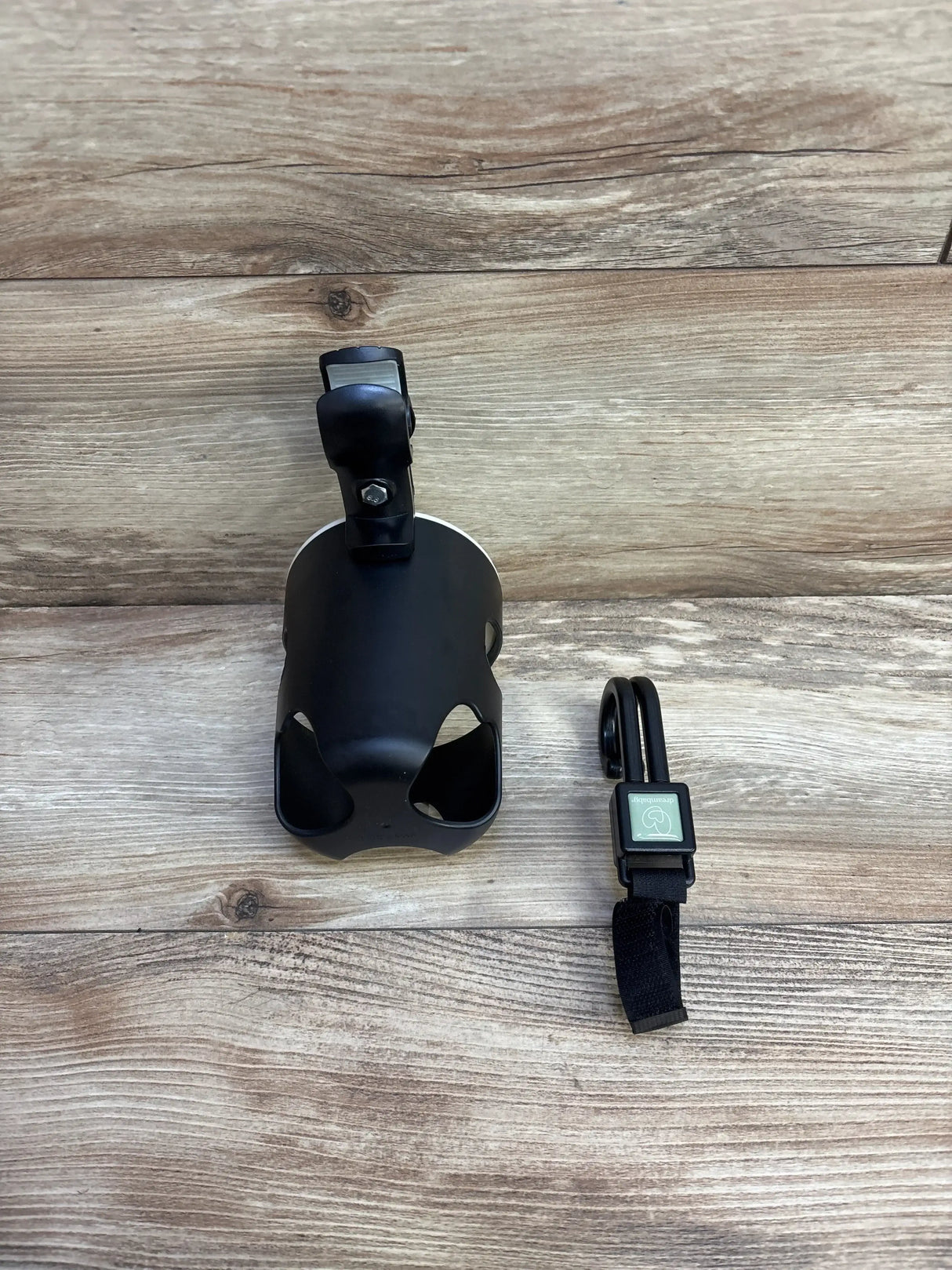 Flat lay of Dreambaby 2pc stroller accessories in black showcasing clip-on bottle holder cup and hook with strap on wood plank floor