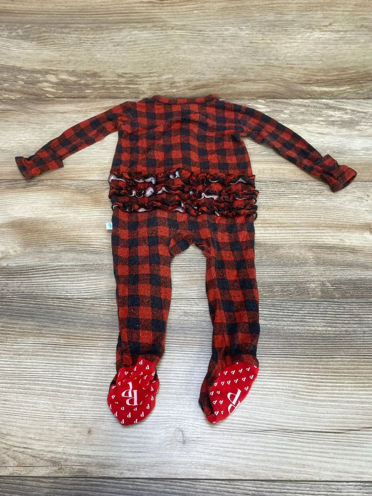 Flat lay of Posh P buffalo-check ruffle footie showing back ruffle detail and built-in red footies on wood background