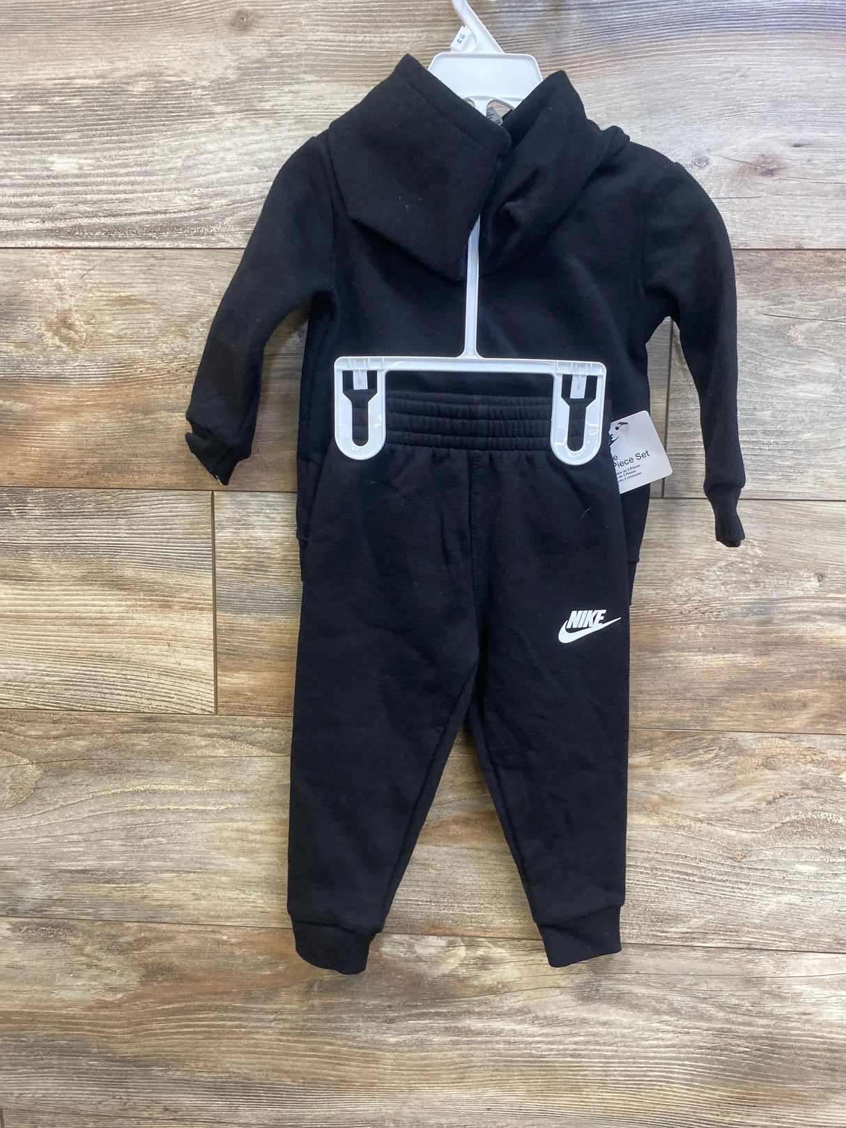 Front view of black Nike toddler hoodie and joggers showing white Nike swoosh and elastic waistband on wooden floor