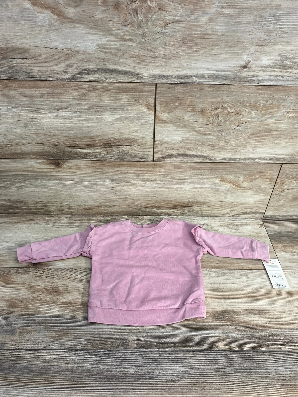 Flat lay of pink Cat & Jack baby sweatshirt (3–6 months) showing long sleeves and shoulder ruffle detail on wood background with tag attached