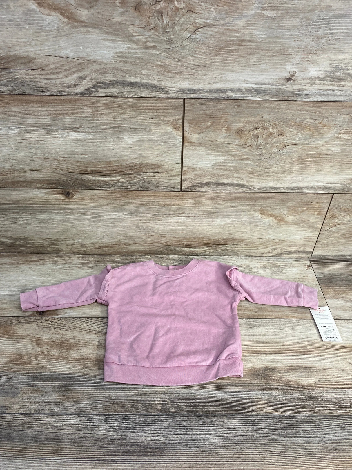 Flat lay of pink Cat & Jack baby sweatshirt (3–6 months) showing long sleeves and shoulder ruffle detail on wood background with tag attached