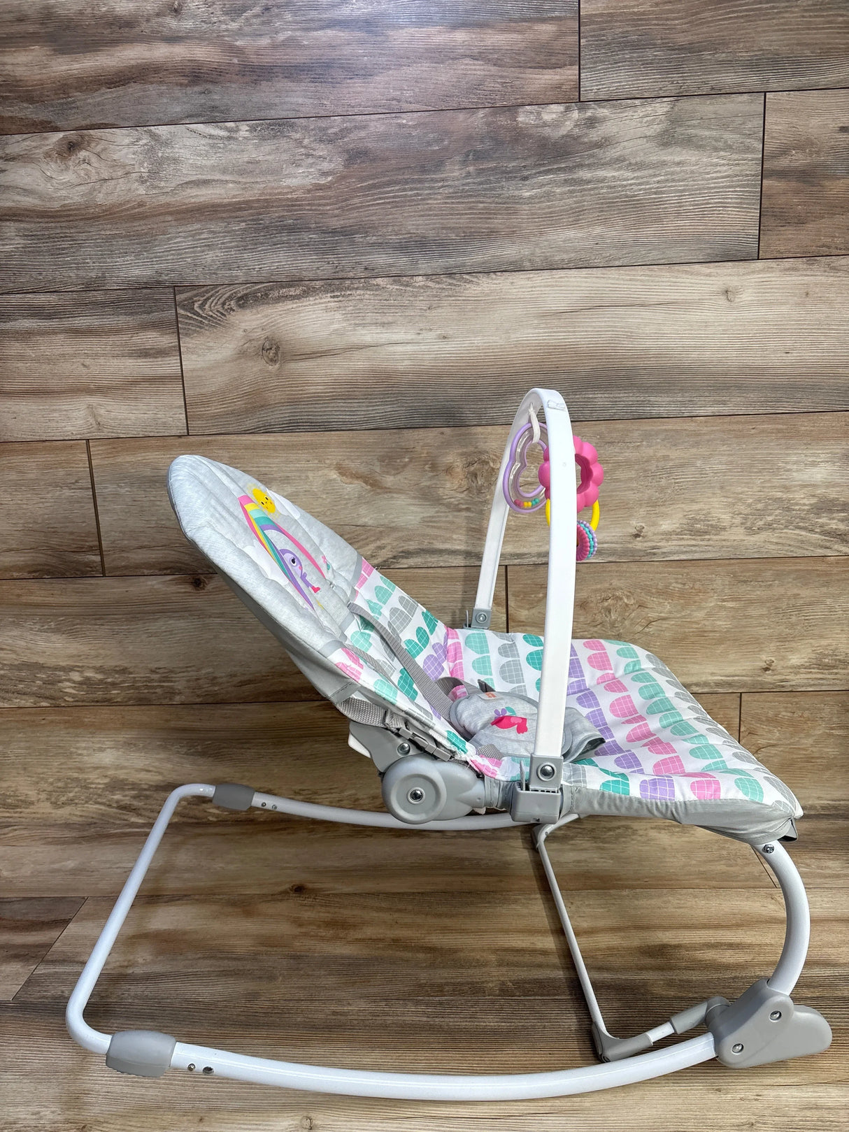 Side view of Bright Starts Rosy Rainbow rocker showing pastel heart-patterned seat, toy bar, white frame on wood floor