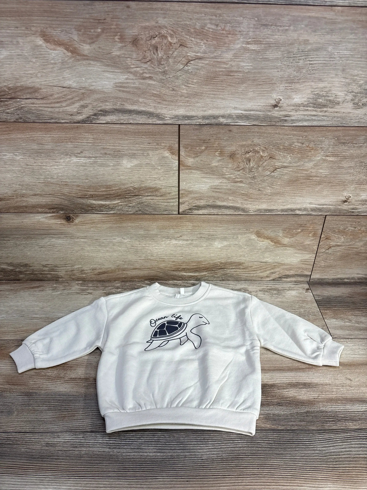 Flat lay of Orchestra long-sleeve pullover sweatshirt, ivory size 12 months, showcasing navy turtle graphic and “Ocean life” text on wooden floor
