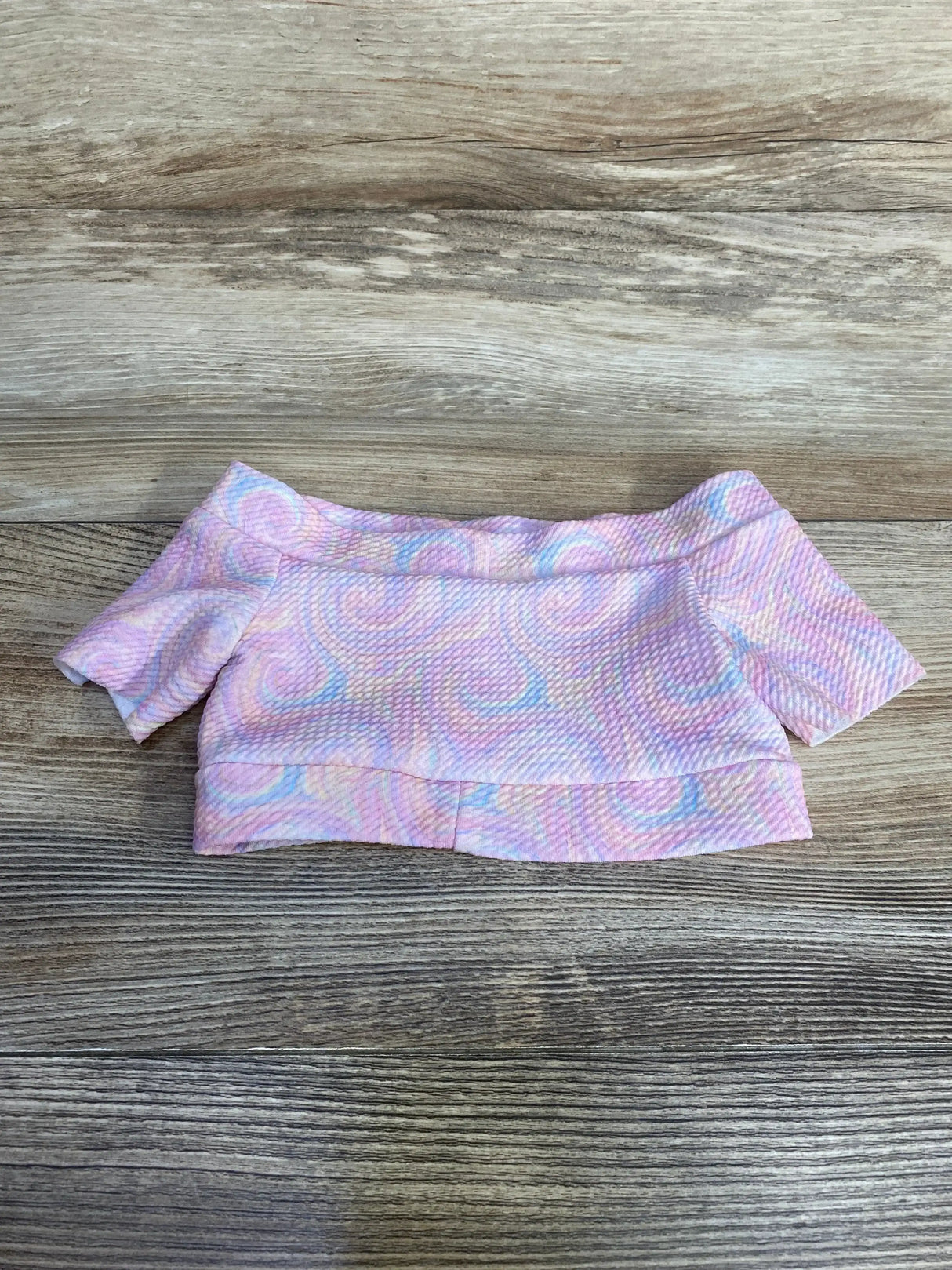 Flat-lay of pink baby cropped top with off-the-shoulder neckline, short sleeves and pastel swirl texture on wood background