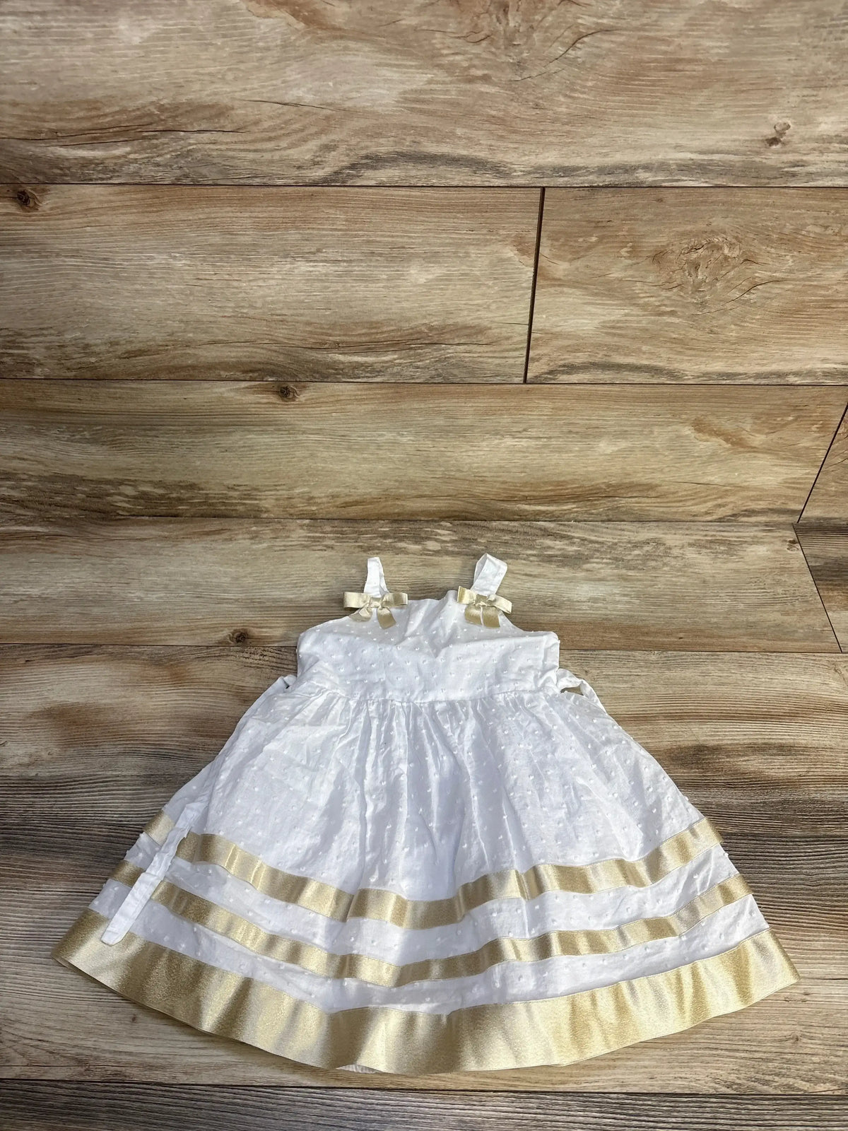 Top down view of girls Bonnie Jean white Swiss dot dress size 2T with gold ribbon hem stripes, shoulder bows, and gathered skirt on wood floor