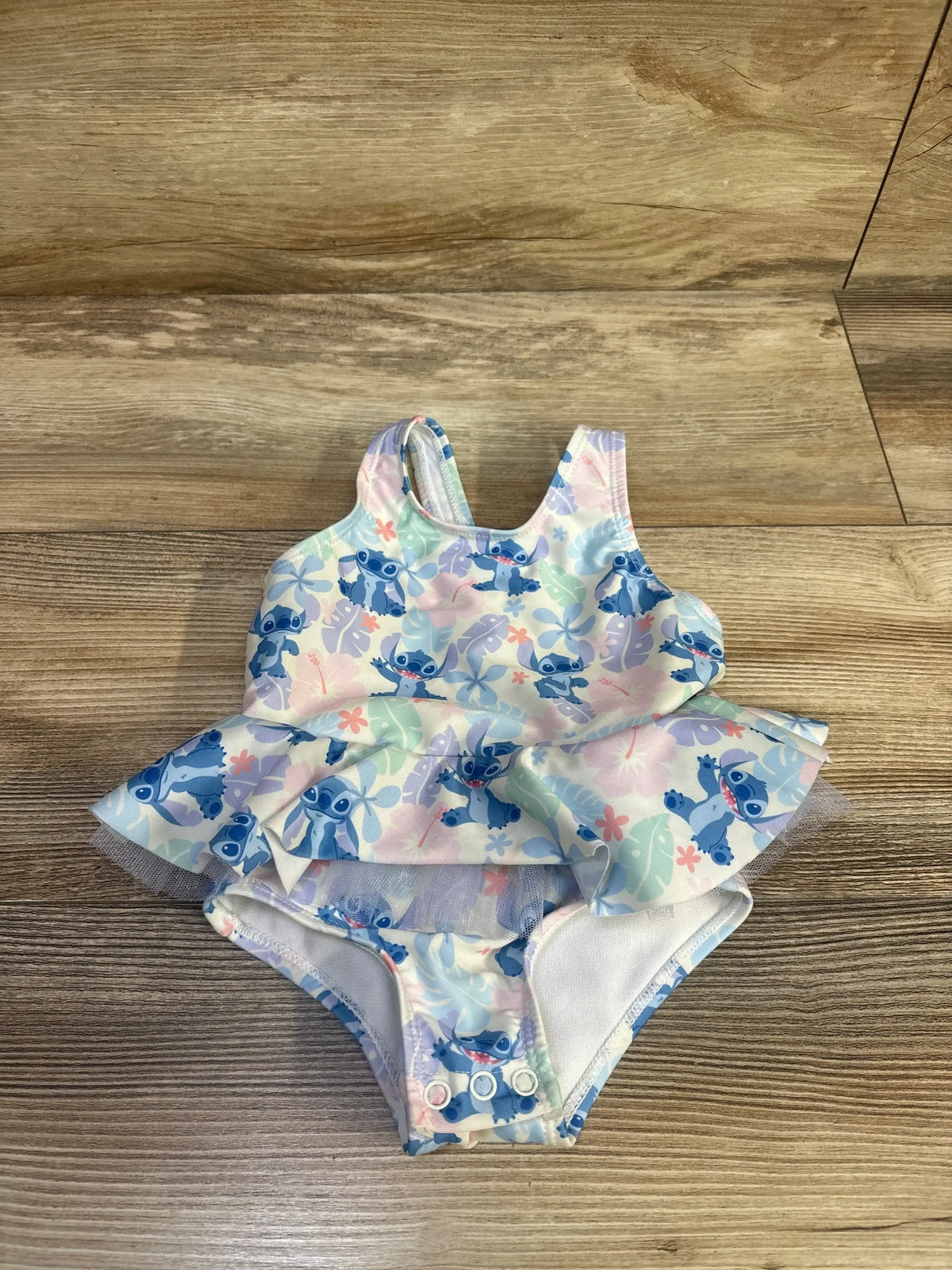 Flat-lay front of baby one-piece swimsuit with Stitch print, tutu ruffle and snap crotch on wood floor