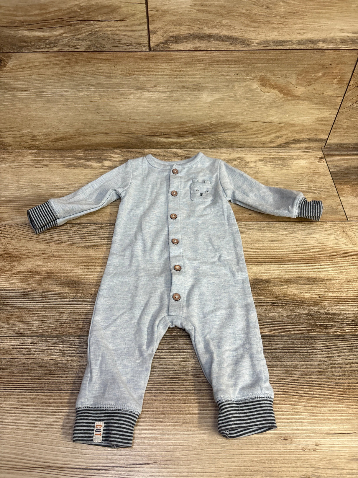 Light gray baby romper with wooden buttons, striped cuffs, and bear face pocket on wooden floor