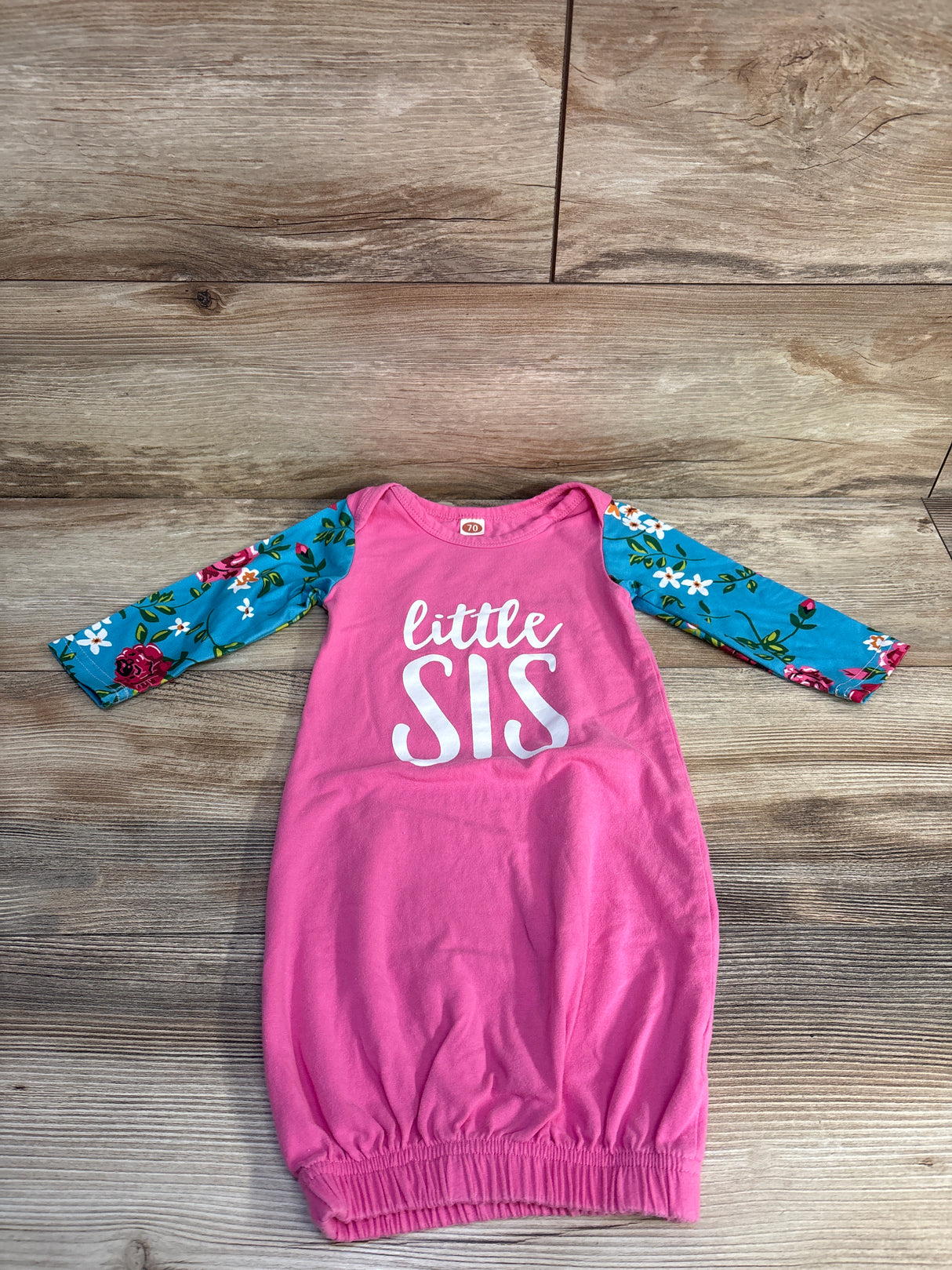 Pink baby gown with 'little SIS' text and blue floral long sleeves on wood background