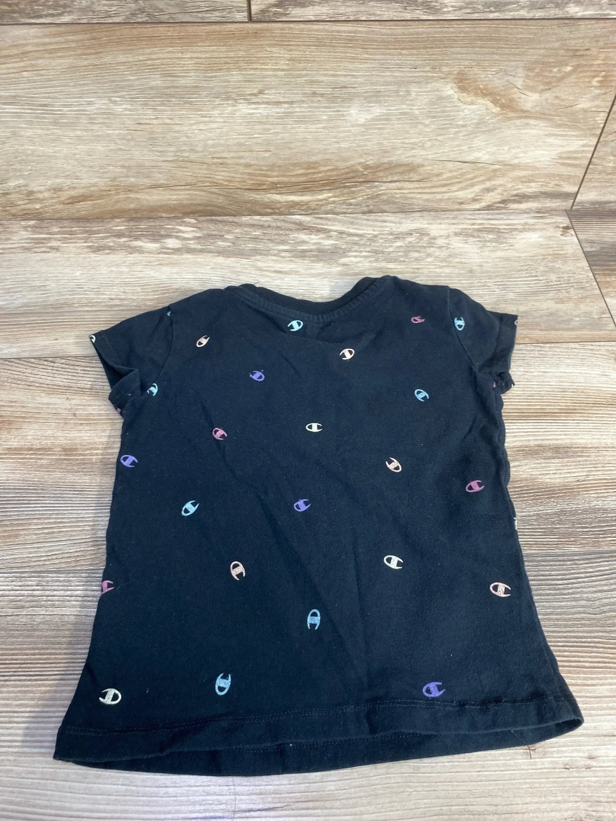 Flat lay of back of black Champion toddler t-shirt showing scattered small multicolored 'C' logos on wood background