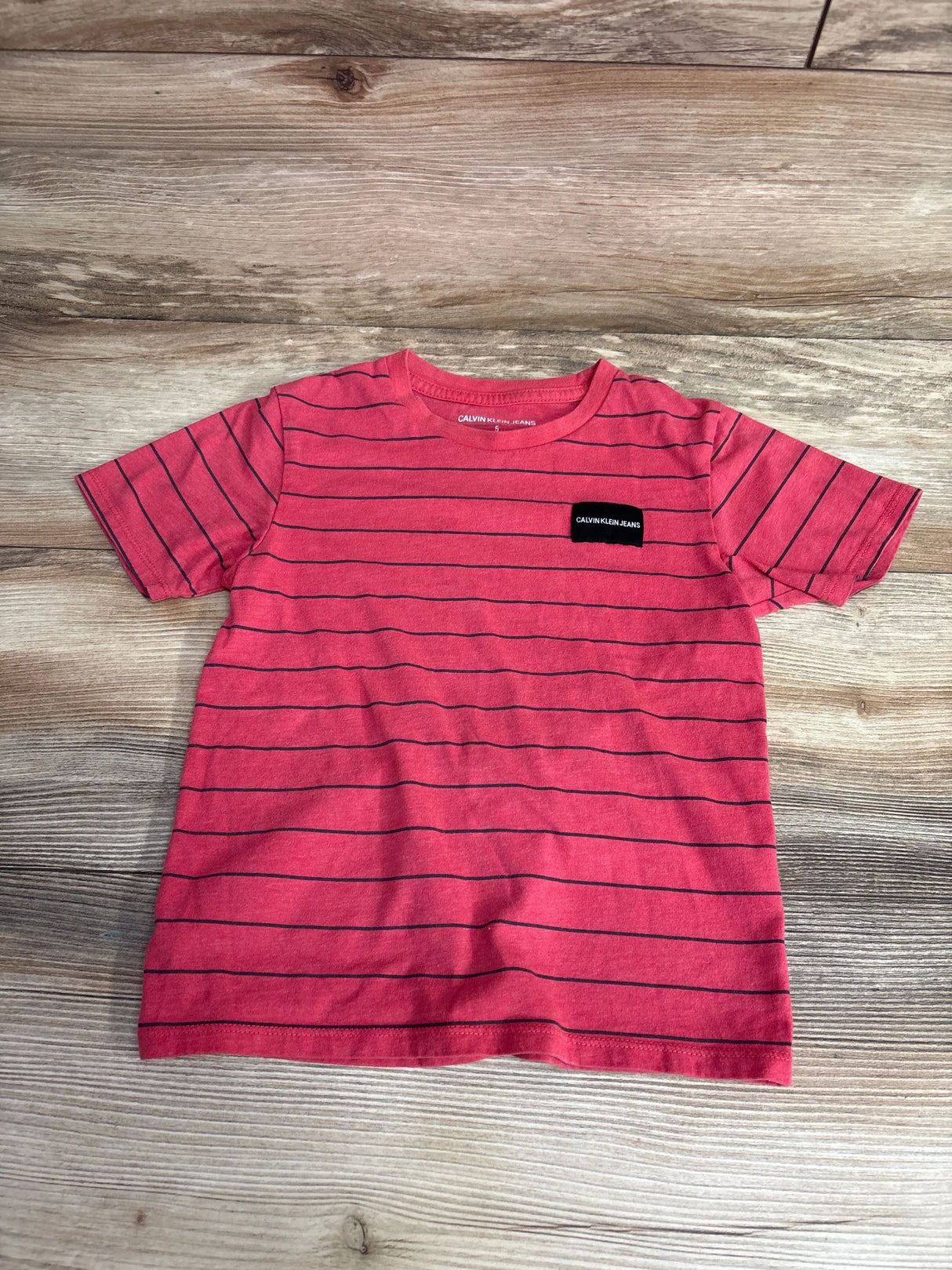 Flat lay of red Calvin Klein short sleeve toddler t-shirt size 5T showing thin black horizontal stripes and small chest logo on wood background