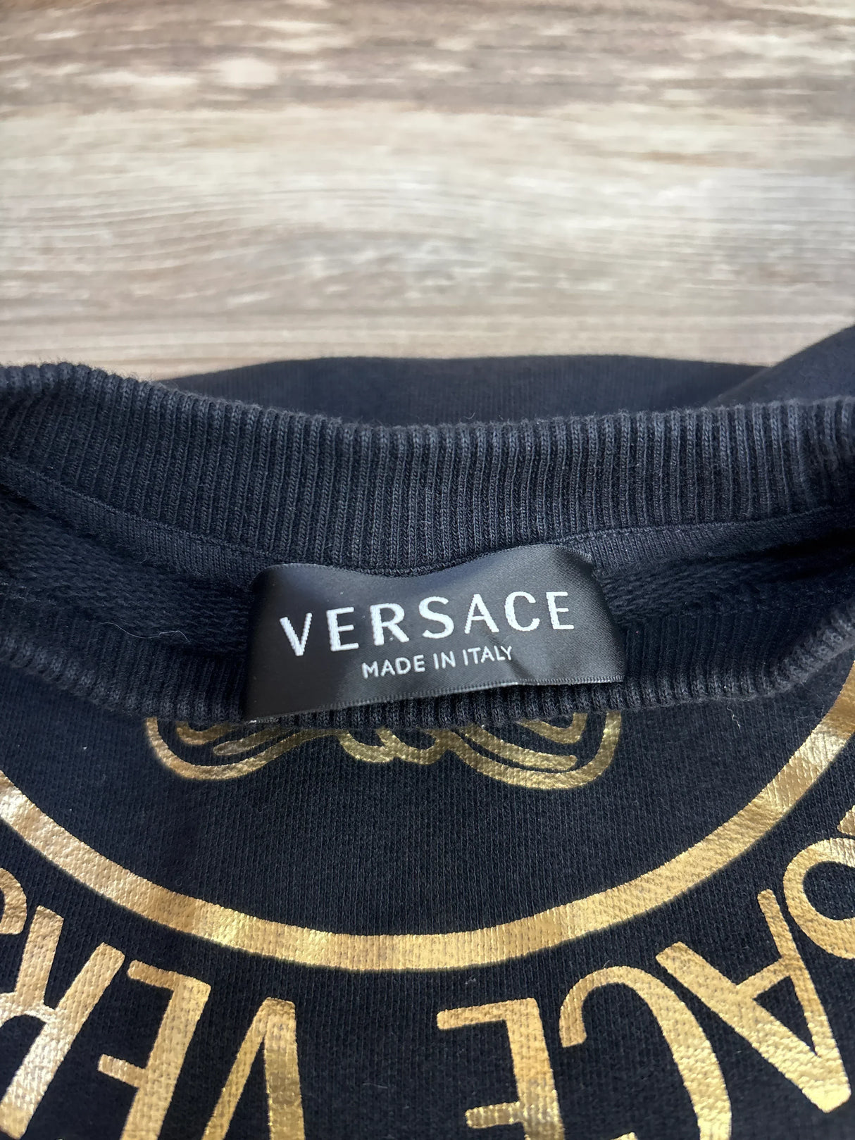 Close-up of Versace black 4T sweatshirt tunic showing neck label VERSACE MADE IN ITALY and gold Medusa print on wooden floor