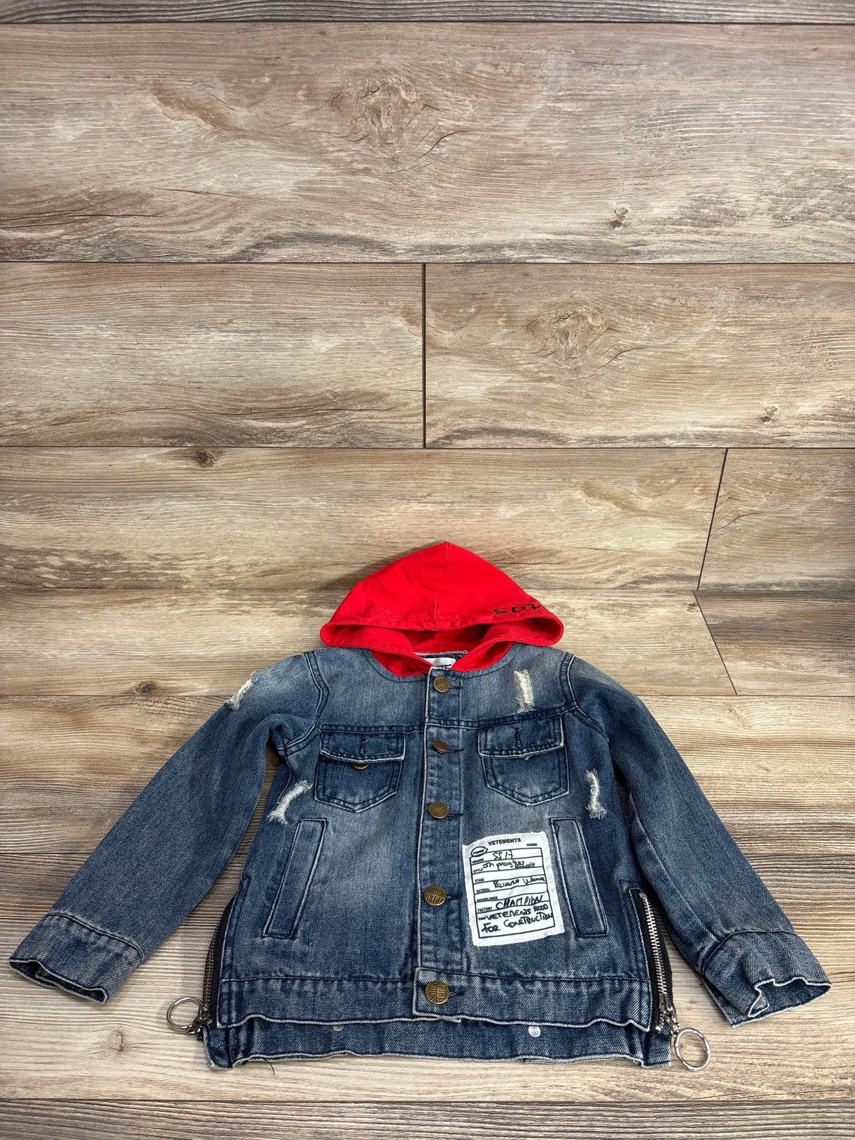 Kids denim jacket with red hood, front pockets, patch and zipper accents, on wood floor