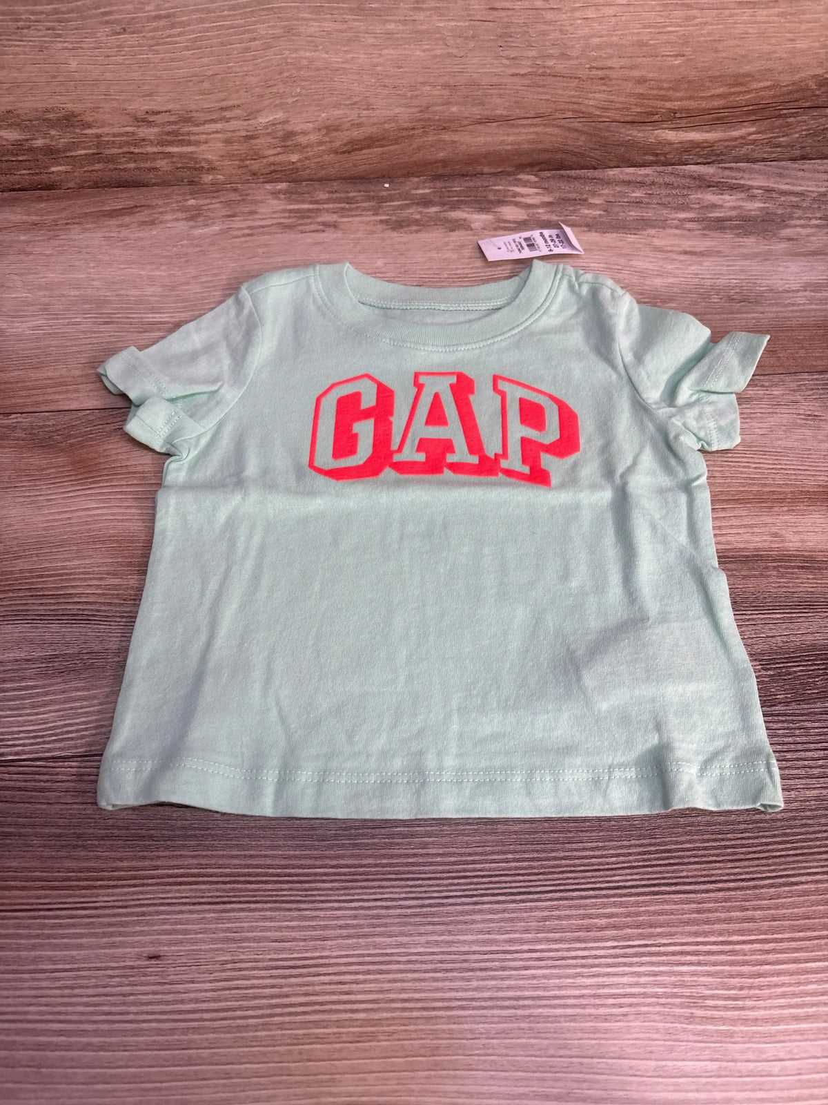 Front view of Baby Gap short-sleeve t-shirt showcasing bright pink GAP logo on mint green fabric with wood-grain background
