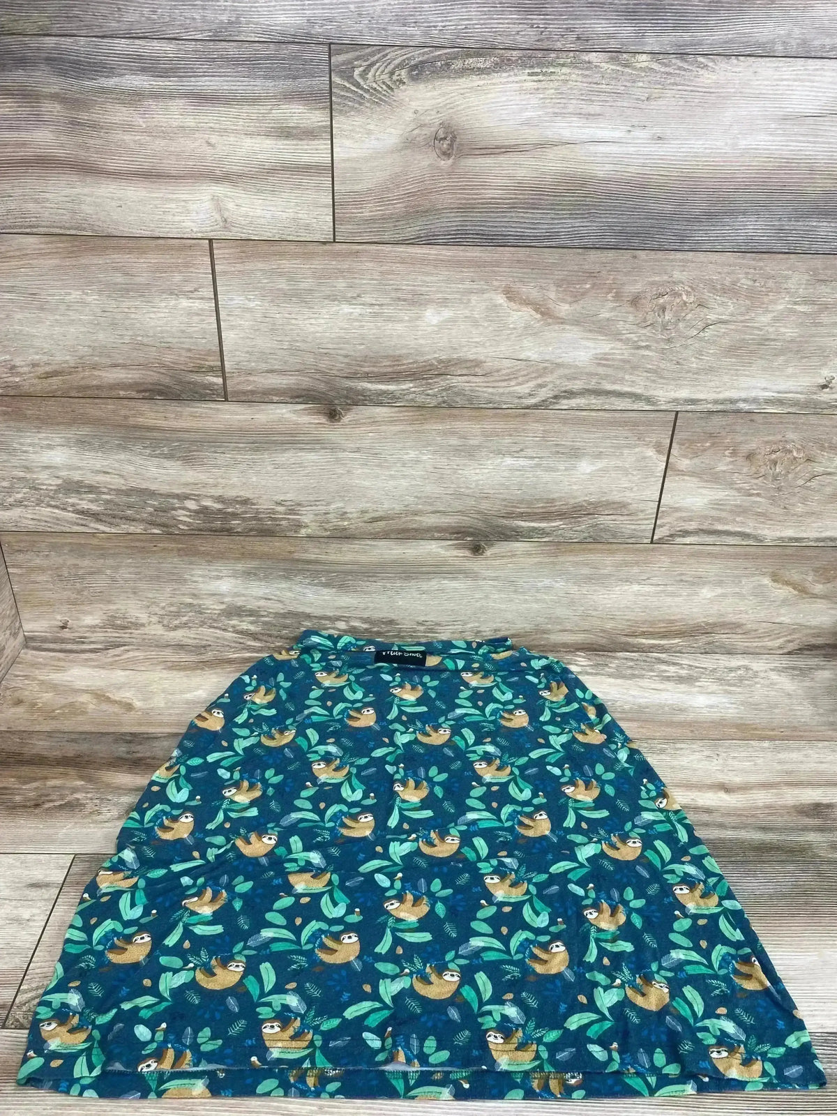 Flat lay of Milk Snob car seat and nursing cover showing green sloth print on teal fabric with wood floor background