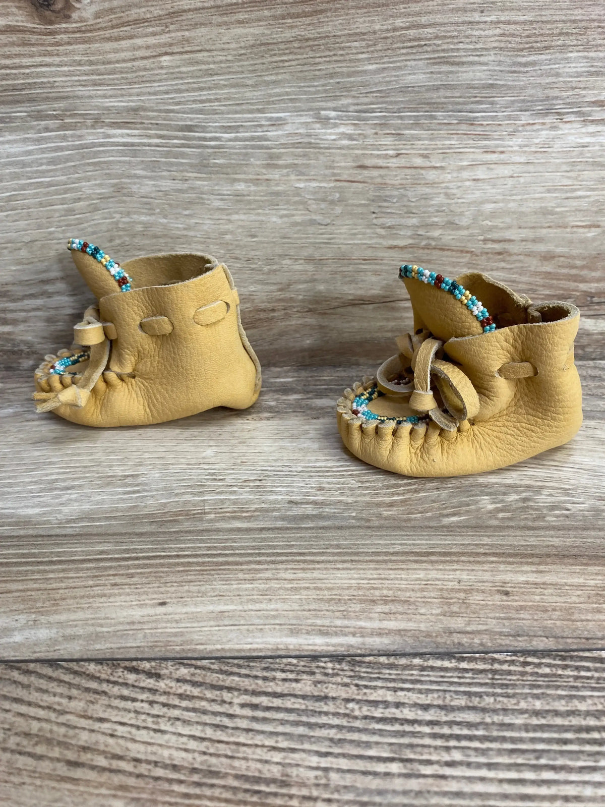 Front-side view of tan leather baby moccasin booties (size 2C) with turquoise-and-white beaded trim, fringe stitching and bow laces on wood background