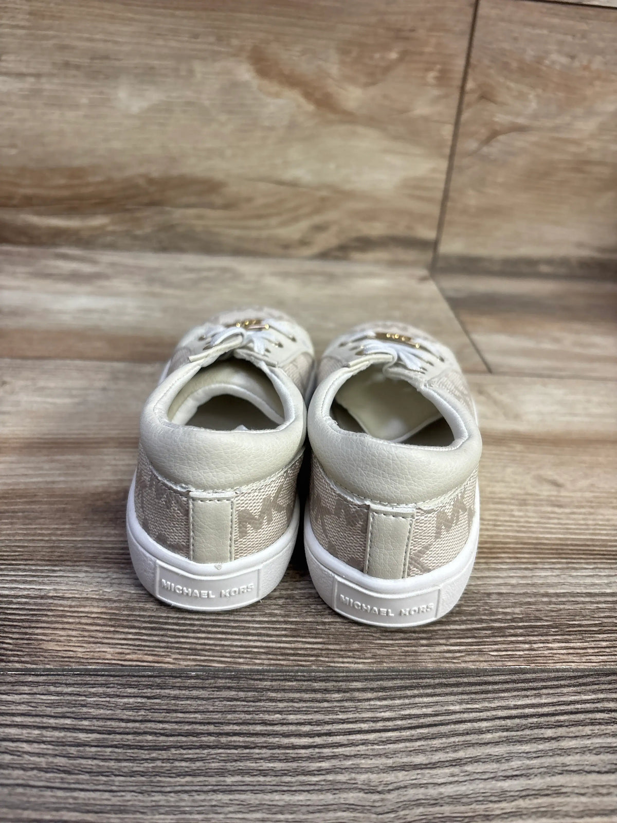 Rear view of Michael Kors logo slip-on sneakers in beige showcasing patterned heel panels and white soles on wood flooring