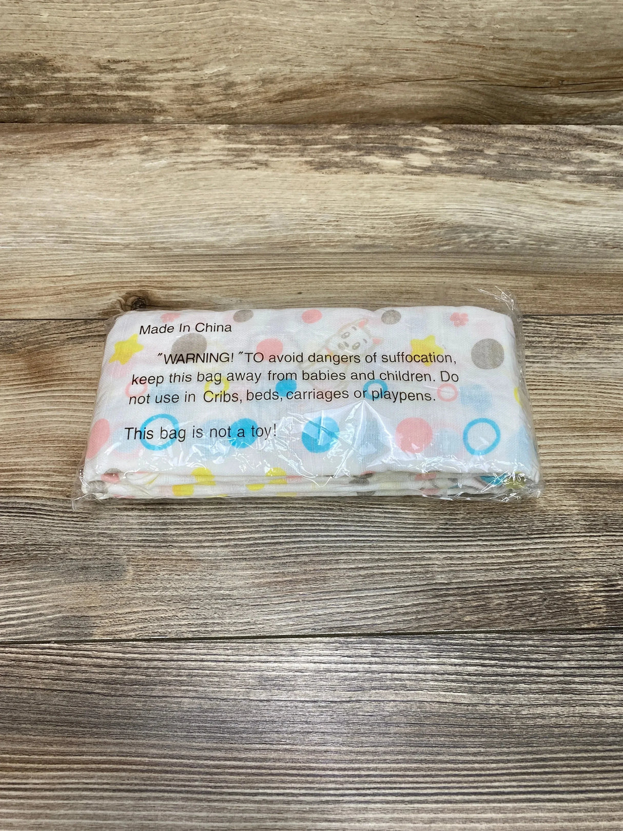 Top view of folded Cucumberries cotton muslin swaddle in clear plastic showing multicolor polka dots and paws on wood background