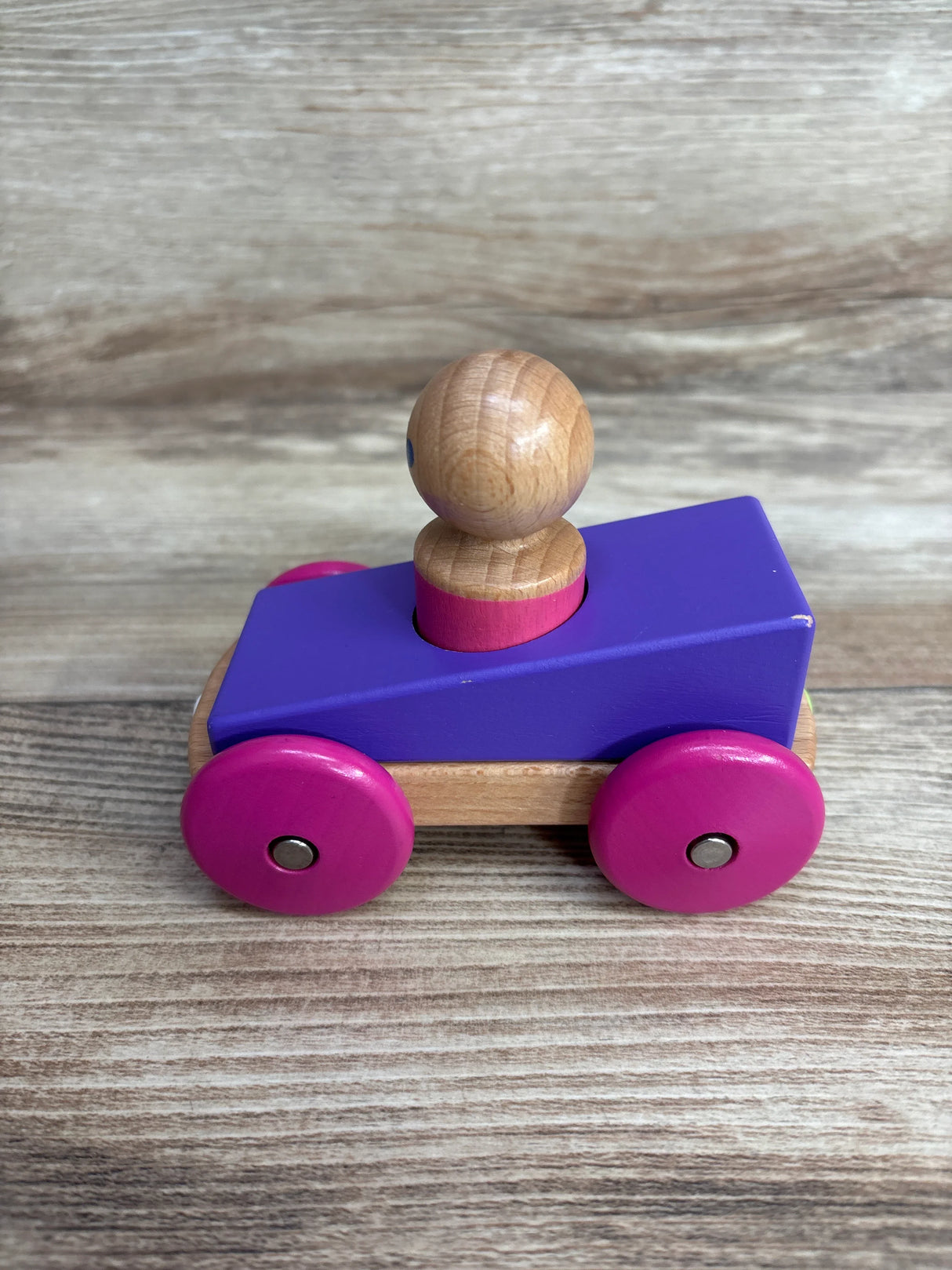 Wooden toy car with purple body, pink wheels, and natural wood driver figure on wood floor