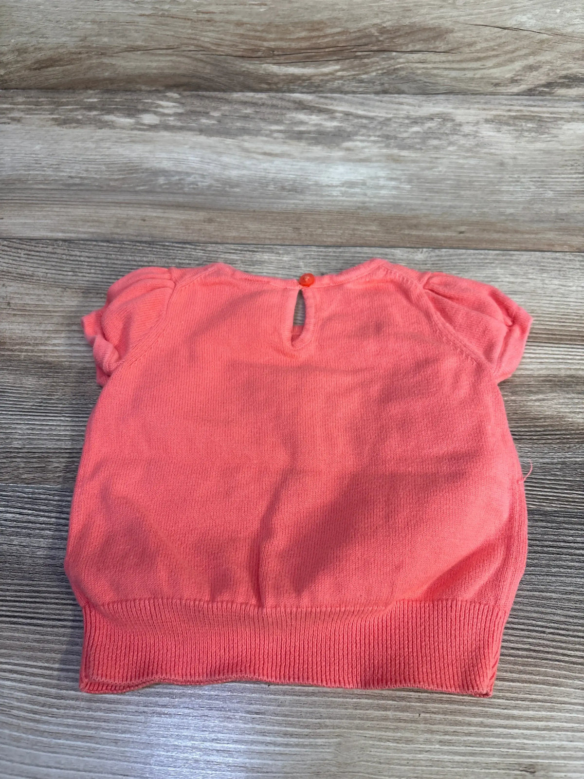 Flat-lay of coral short-sleeve knit sweater showing back keyhole button, puff sleeves and ribbed hem on light wood