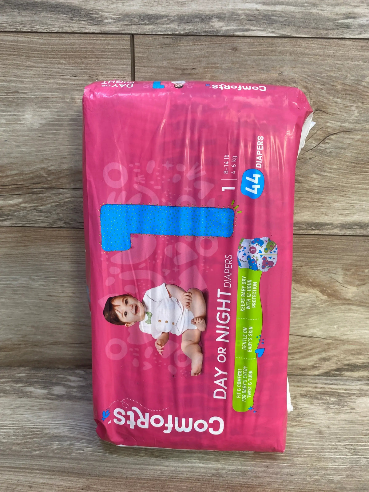 Top view of Comforts size 1 diaper pack, pink packaging with baby image, Day or Night label, 44 count, 8 to 14 lb on wood