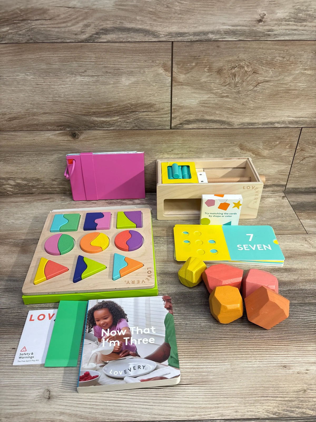 Overhead view of Lovevery Free Spirit kit with shape puzzle, storage box, counting cards and chunky blocks on wood backdrop