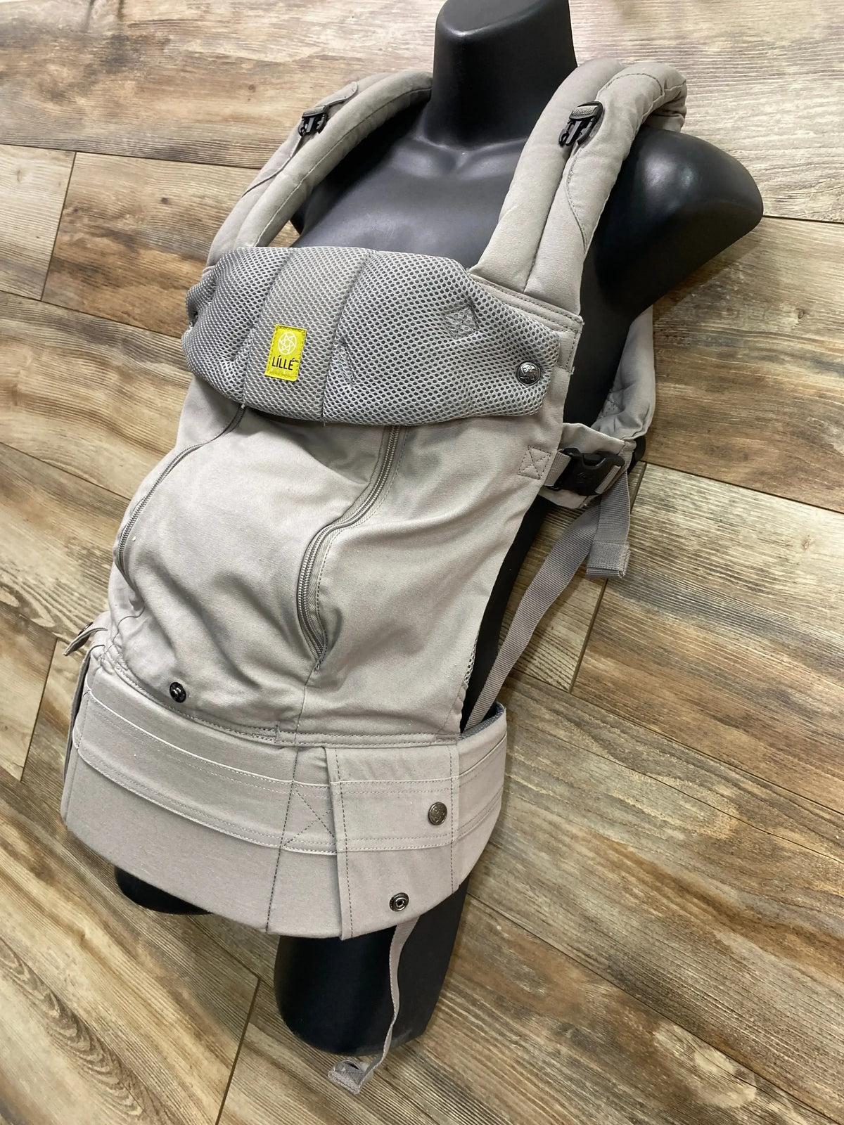 Angled front of Lille Baby carrier in stone grey showing mesh panel and padded straps on black mannequin over wood floor