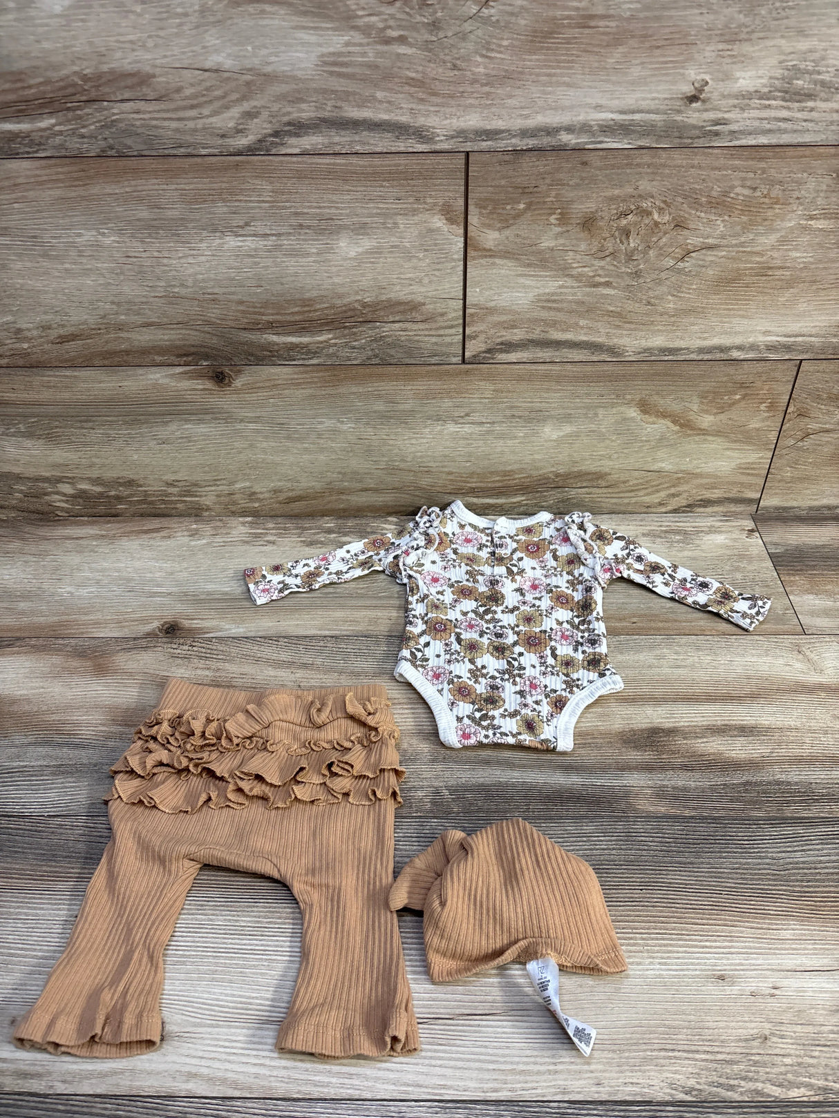 Flat lay of 3-piece baby set: white floral long-sleeve bodysuit with ruffle shoulders, tan ruffle pants and hat on wood