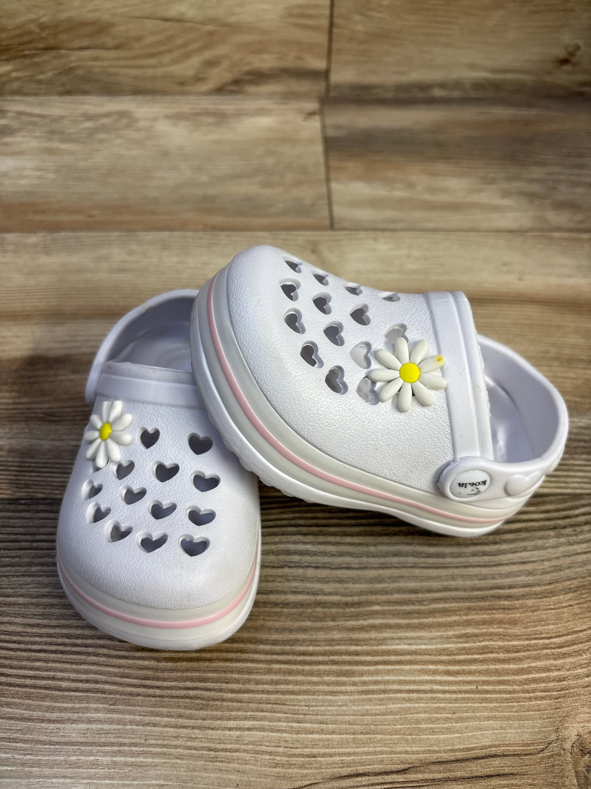 Front and side view of white Koala Kids toddler clogs showing heart cutouts, pink sole stripe, and daisy charm on wood surface