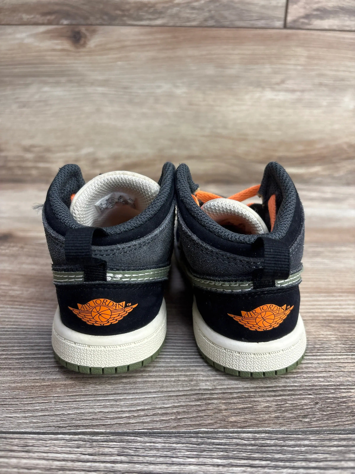 Kids black and grey Air Jordan sneakers with orange logo, rear view, on wood floor