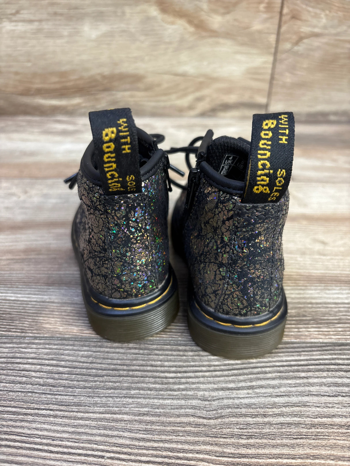 Rear view of Dr. Martens 1460 Crinkle Metallic toddler boots (size 5C) showcasing black crinkle metallic glitter finish and yellow heel loops on wood floor