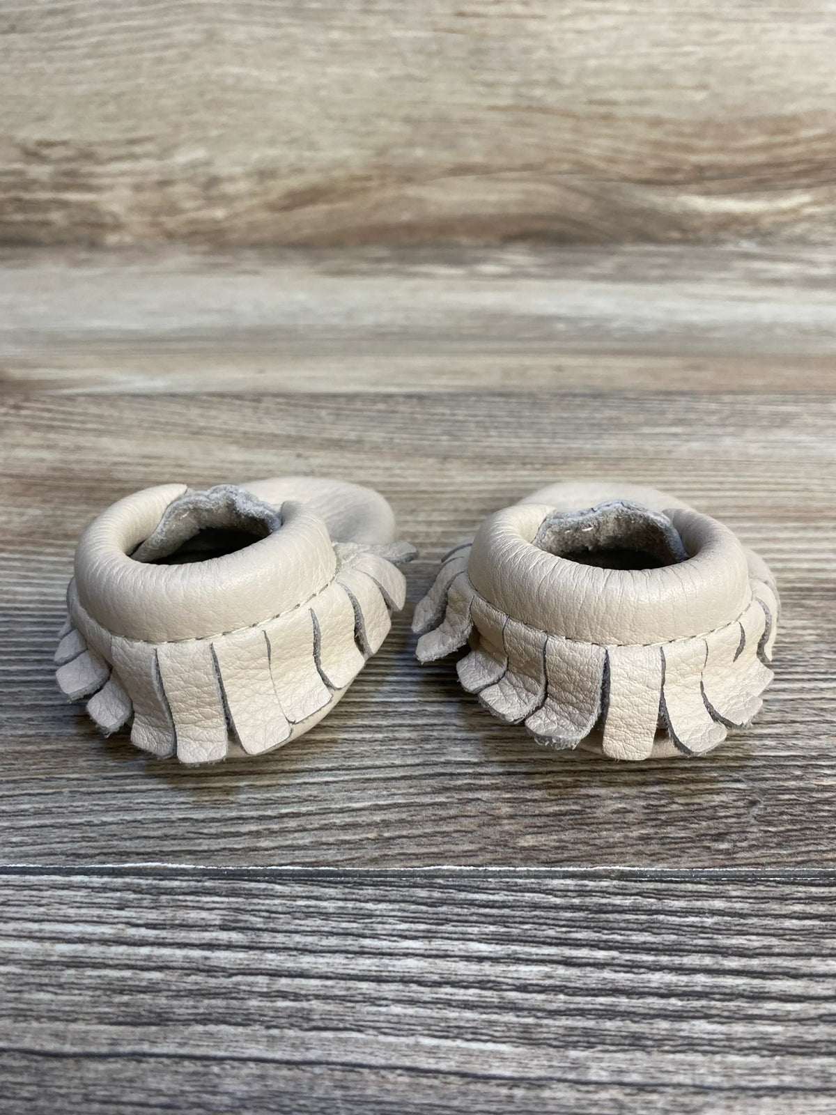 Beige leather baby moccasins with fringe on wooden background, gently used infant footwear