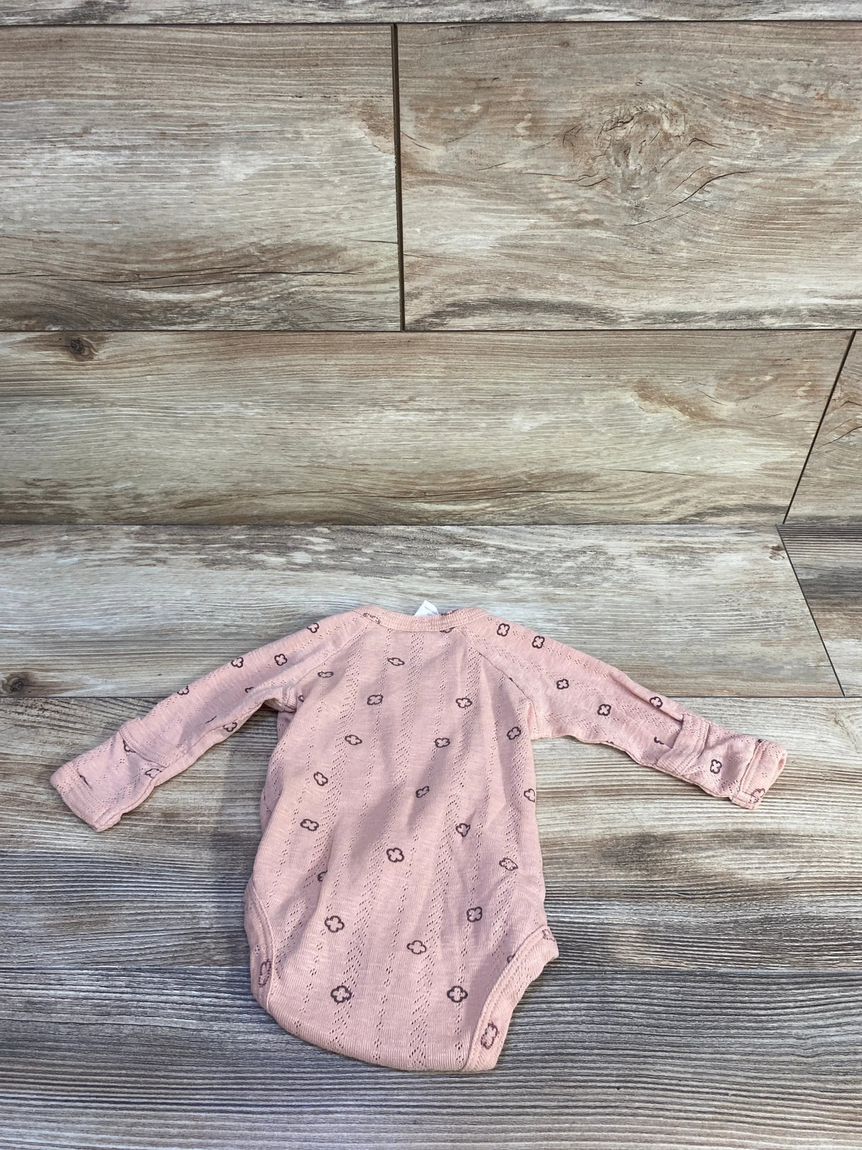 Flat lay of pink Kate Quinn organic kimono newborn bodysuit showcasing long sleeves and small floral eyelet pattern on wood plank background
