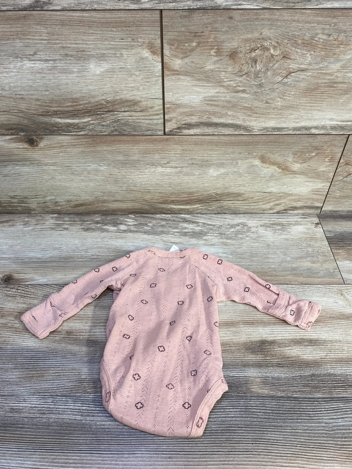 Flat lay of pink Kate Quinn organic kimono newborn bodysuit showcasing long sleeves and small floral eyelet pattern on wood plank background