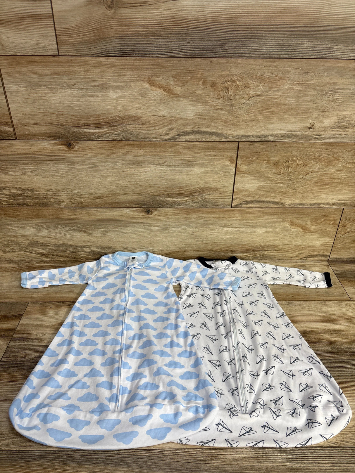 Two baby sleep sacks with zippers on wooden floor, blue clouds and black paper plane patterns