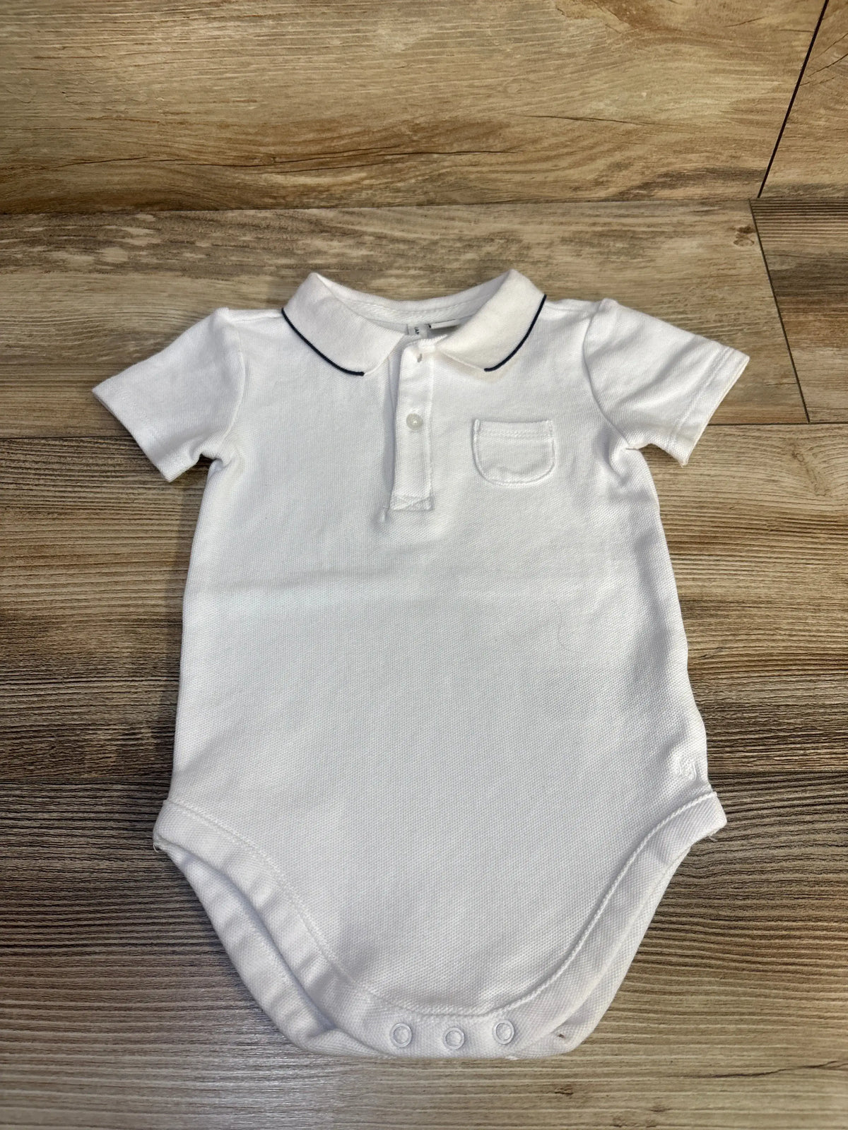 Front view of white Janie & Jack polo bodysuit showcasing navy-trimmed collar, chest pocket, and snap closures on wood flooring