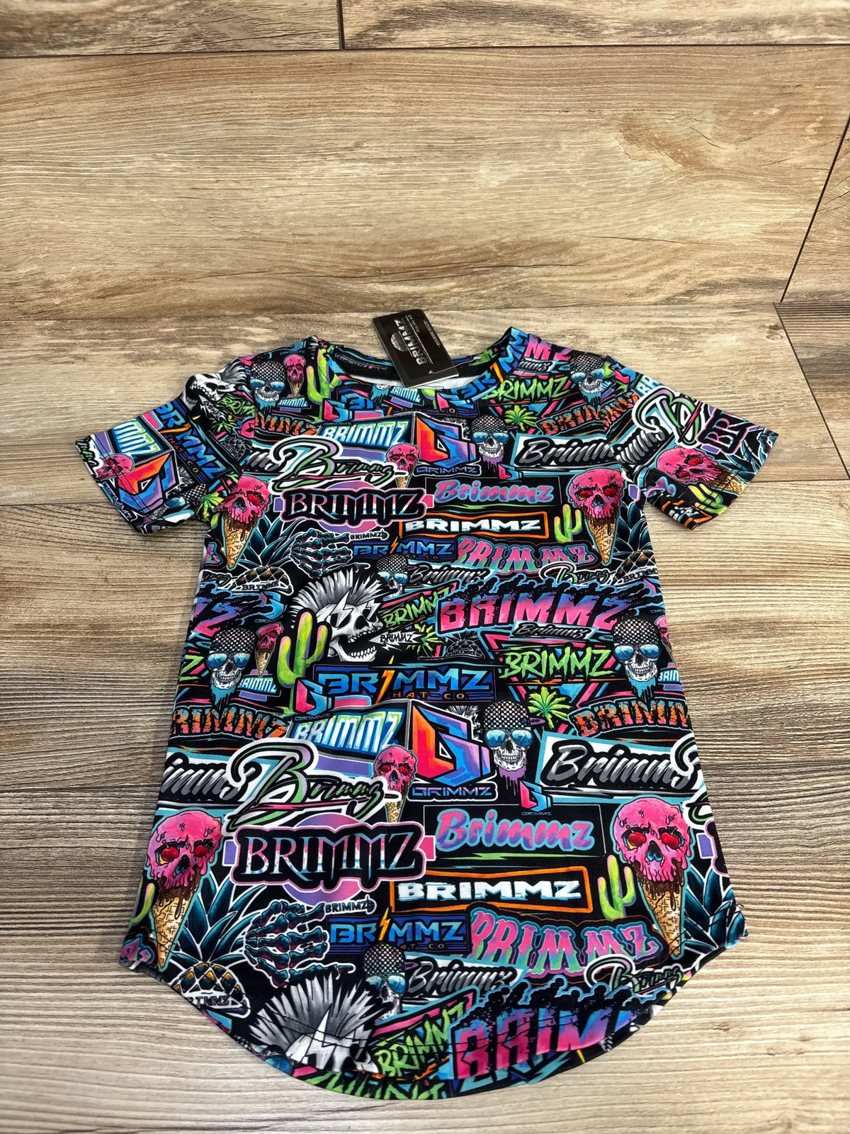 Colorful graphic t-shirt with skulls, cacti, and "Brimmz" text pattern on wood floor