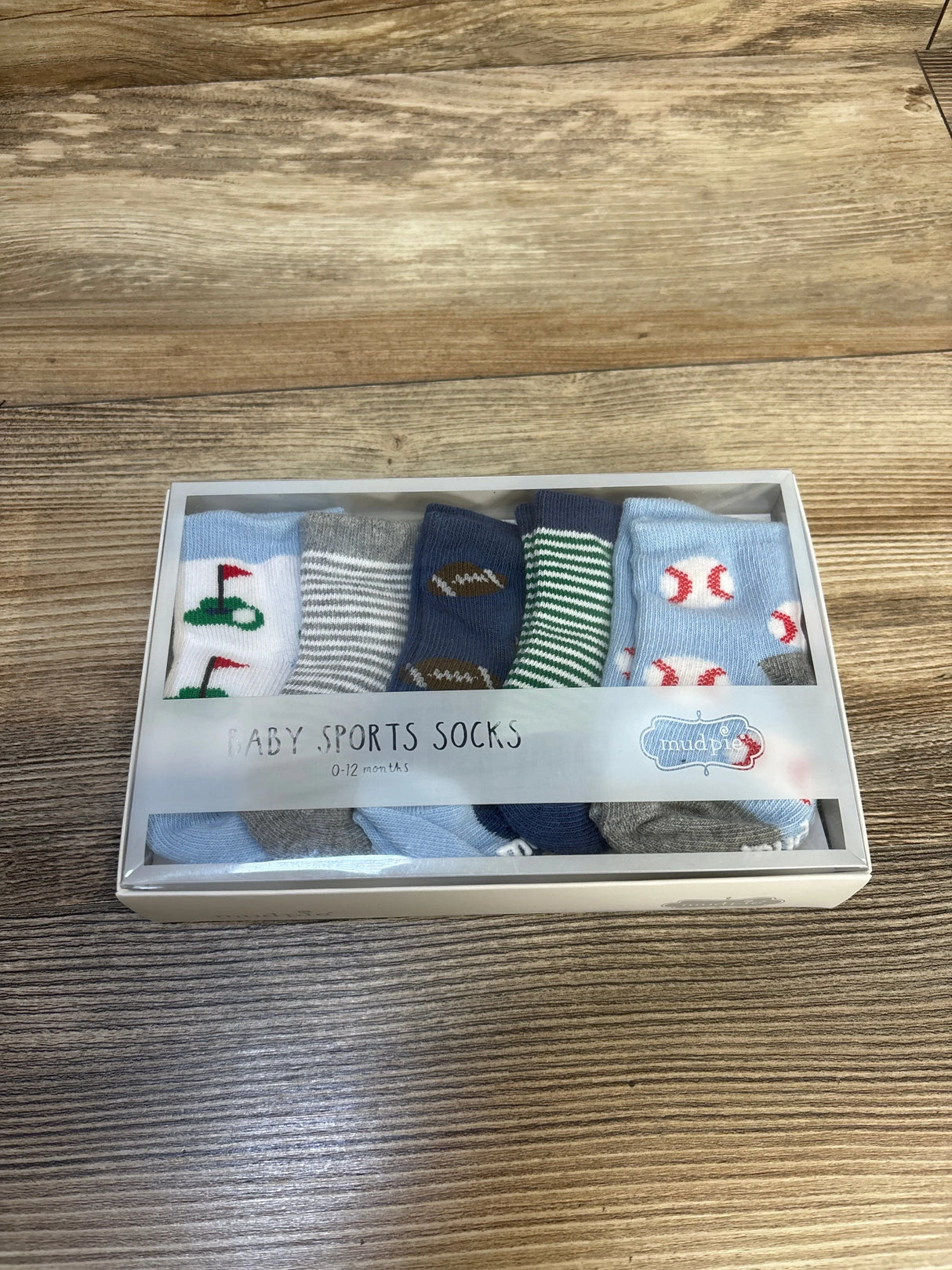 Top-down view of Mud Pie baby sports socks 5-pack showcasing golf, football, and baseball patterns with clear boxed packaging on wooden surface