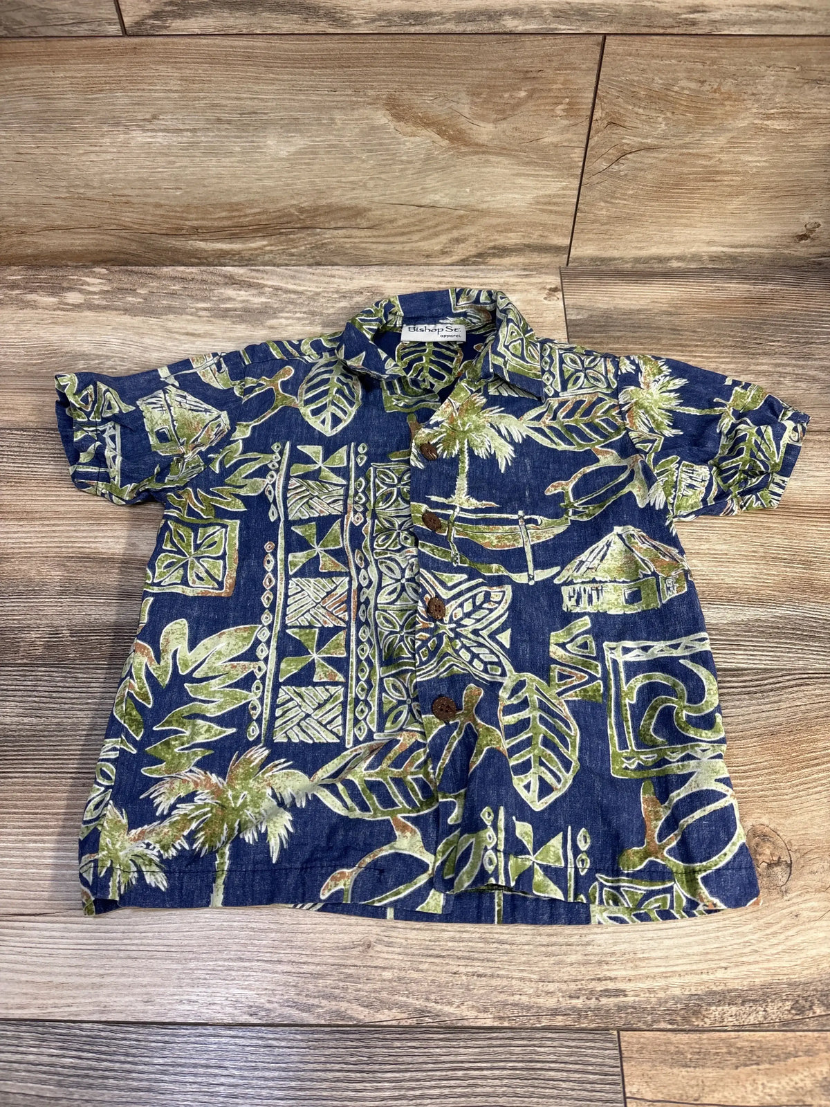 Flat lay of blue Bishop St. Hawaiian short sleeve toddler shirt (size 5T) showcasing green palm and tiki print on a wood background