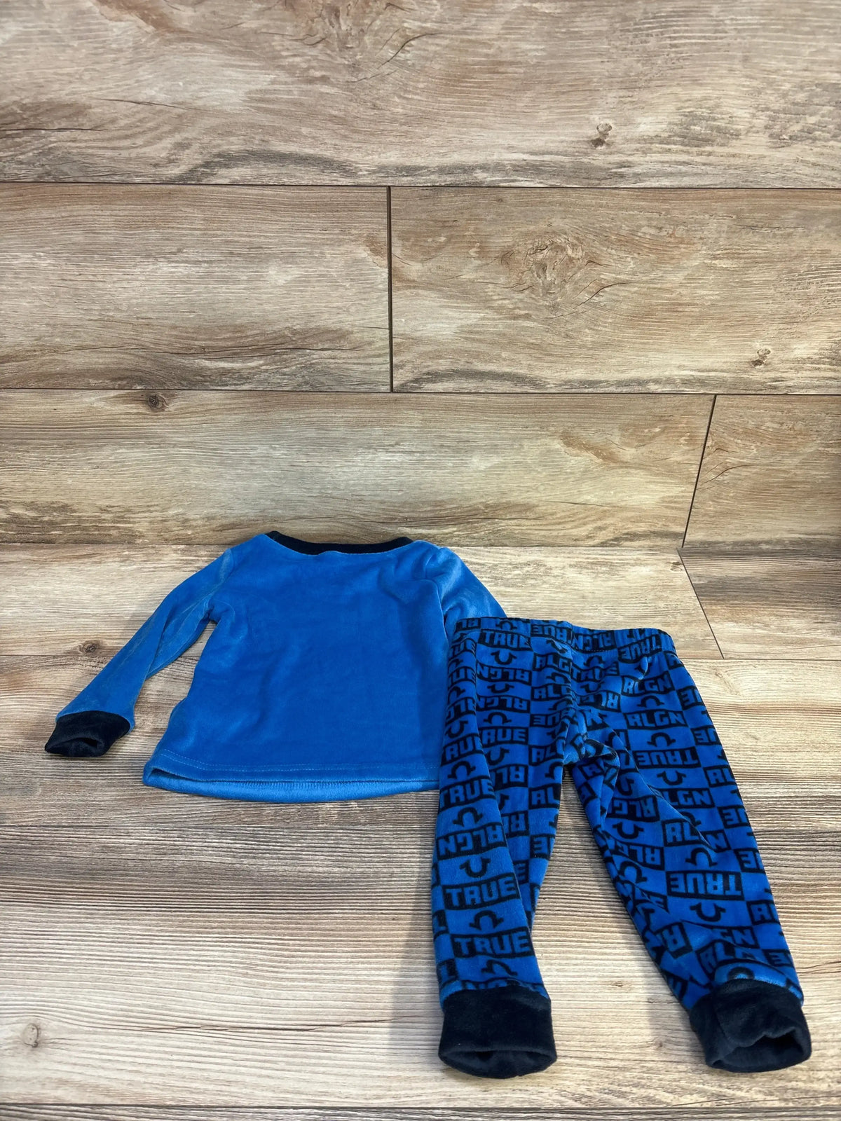 Flat lay of toddler blue True Religion 2-piece fleece pajama set showcasing long-sleeve top and logo-print pants on wood floor