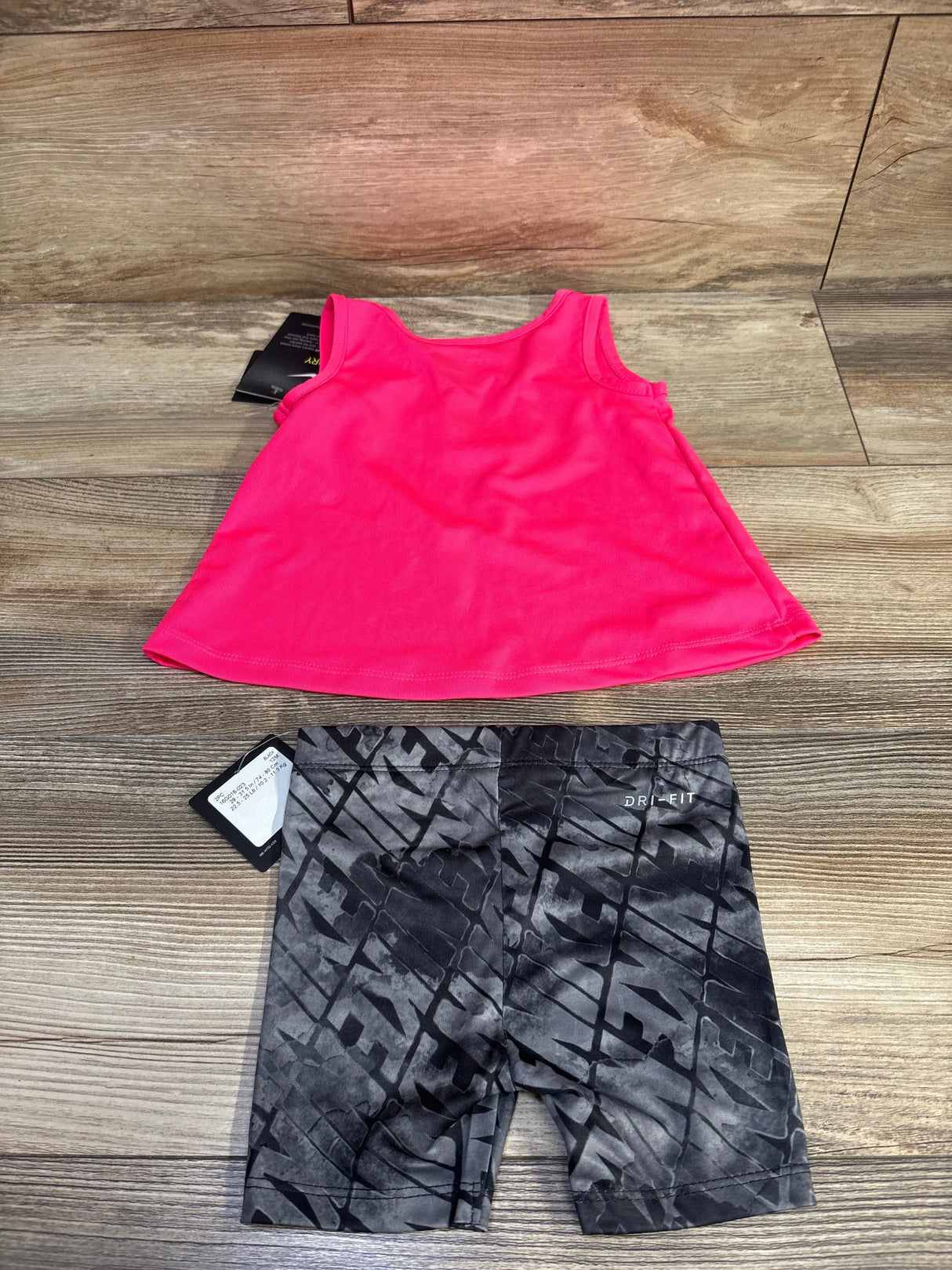Flat-lay of 12M hot pink Nike Dri-FIT tank and gray patterned Dri-FIT shorts, front view with tags on wood background