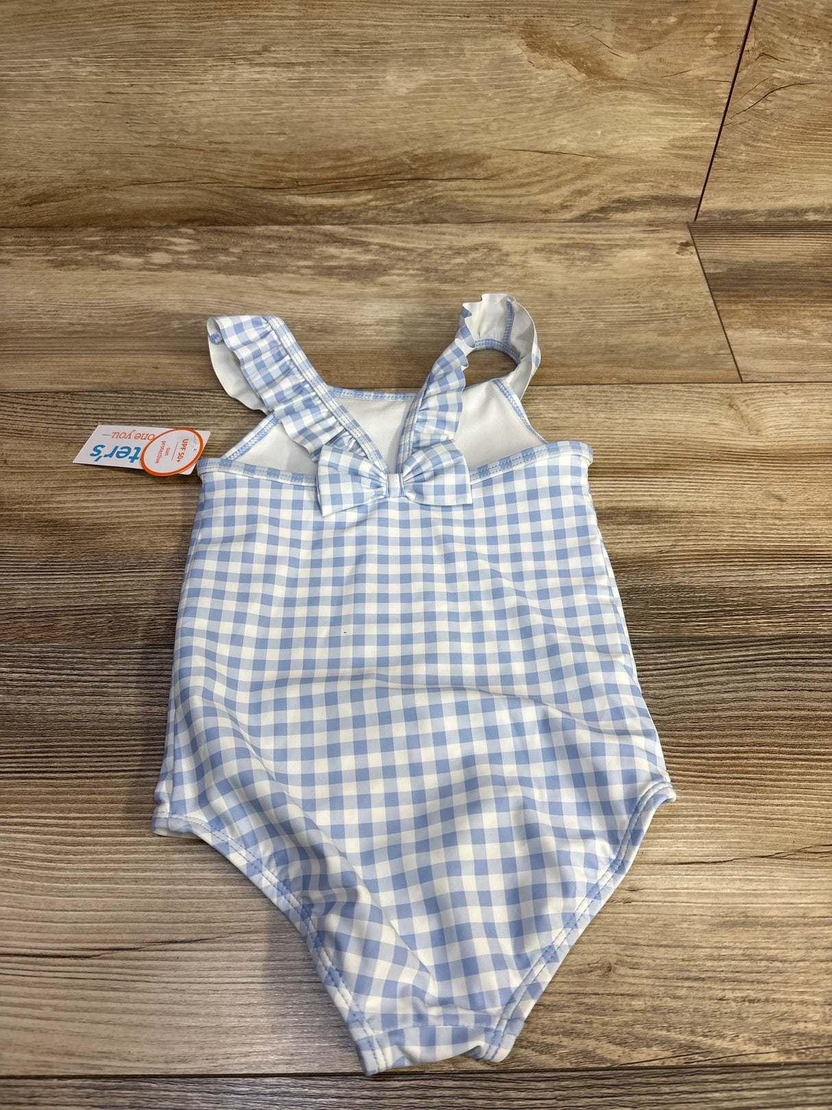 Flat-lay of blue and white gingham one-piece toddler swimsuit showing front bow and ruffled straps on wood background