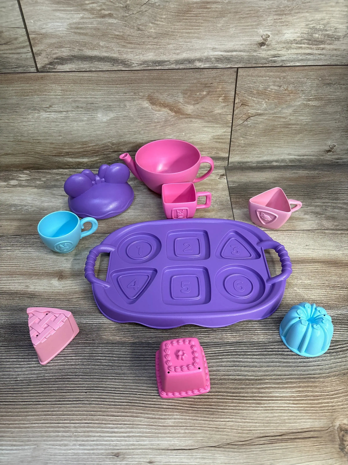 Top-down of Green Toys Minnie Mouse tea party set showing purple numbered tray, pink teapot bowl, cups, and cake molds