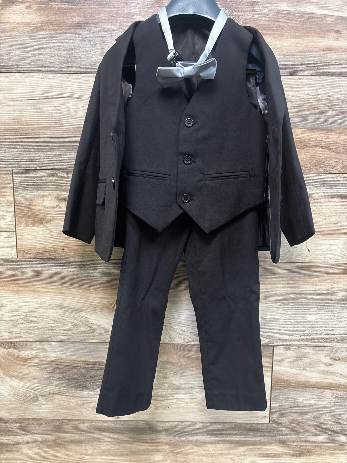 Front view of Boltini black 4-piece suit set showcasing notch lapel blazer, vest, dress pants, and bowtie on a wood floor