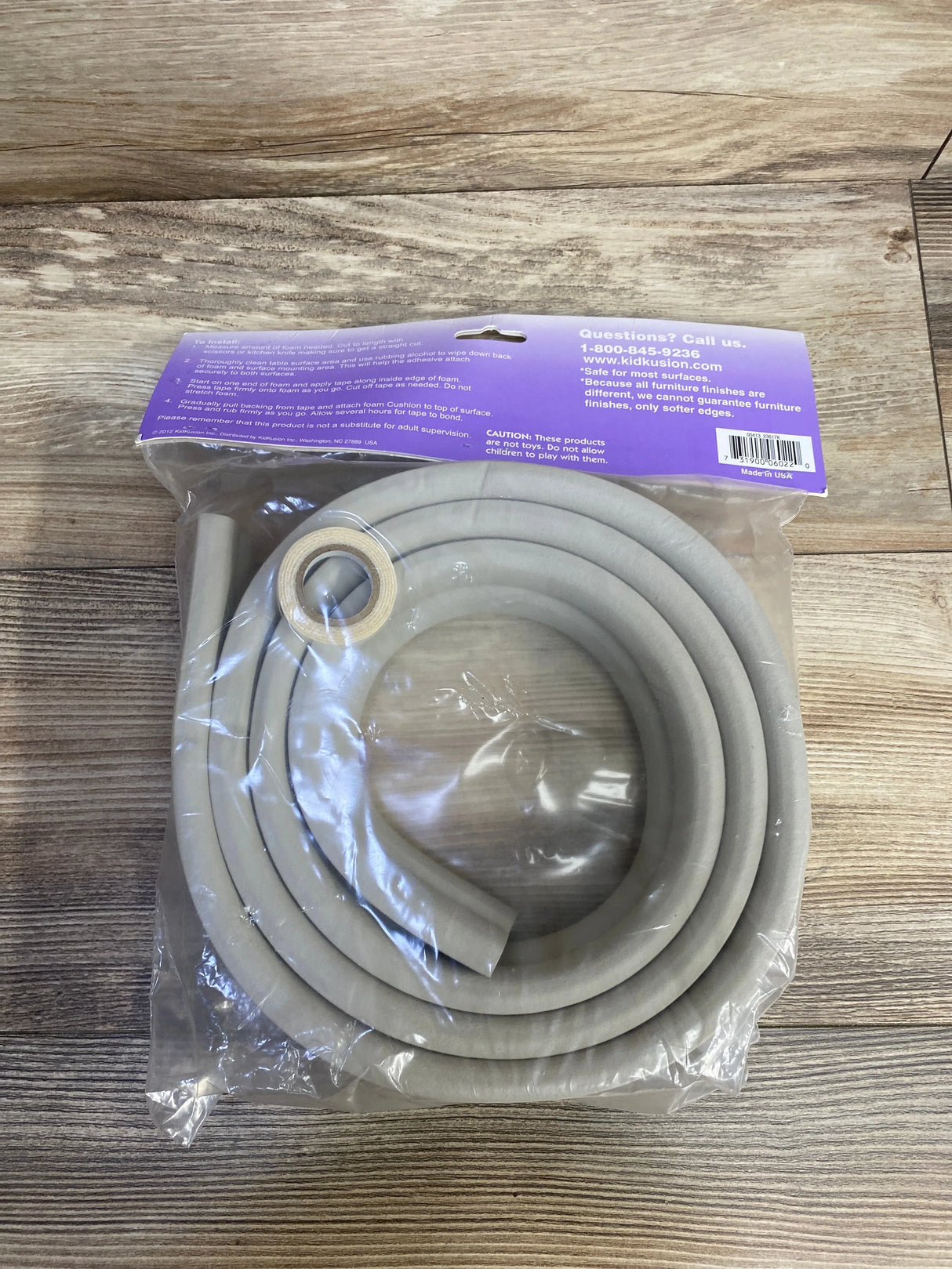 Top-down view of KidKusion Jumbo Edge Cushion in taupe, coiled in original packaging showing thick foam tubing and included adhesive tape on wood background