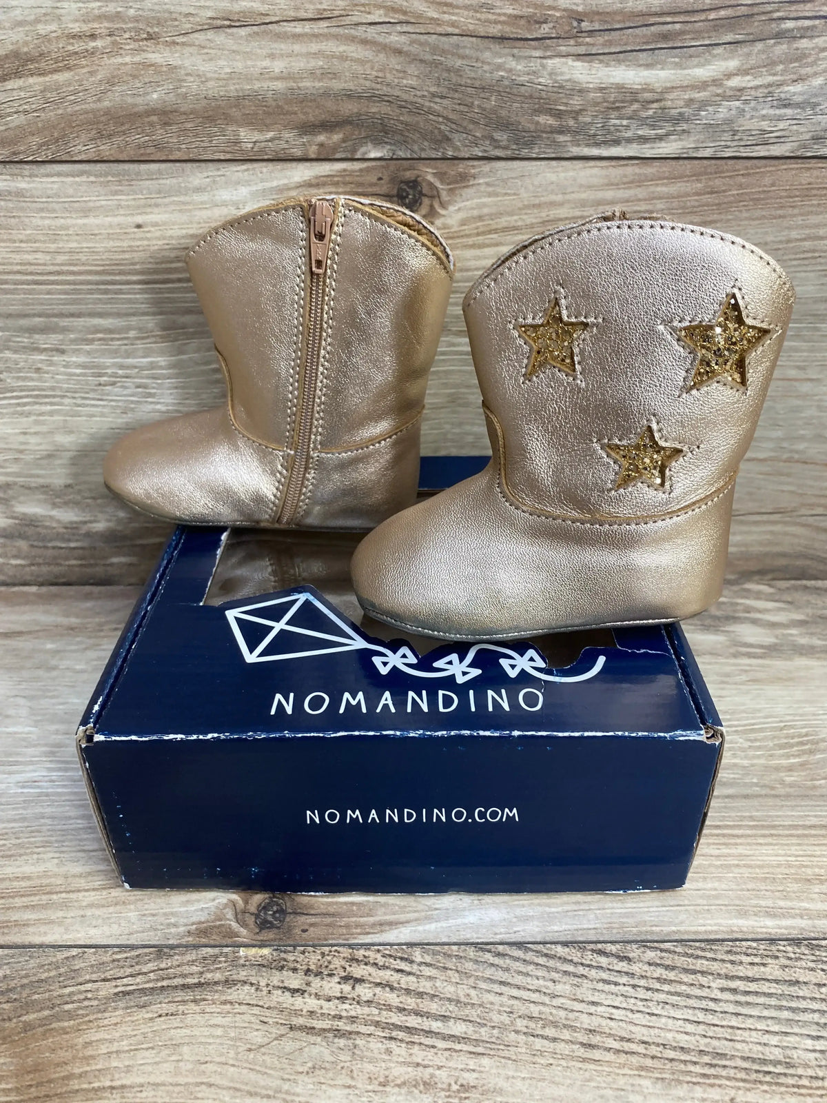 Three-quarter view of Nomandino spark gold baby boots with glitter star inlays and side zipper on navy box, wood background