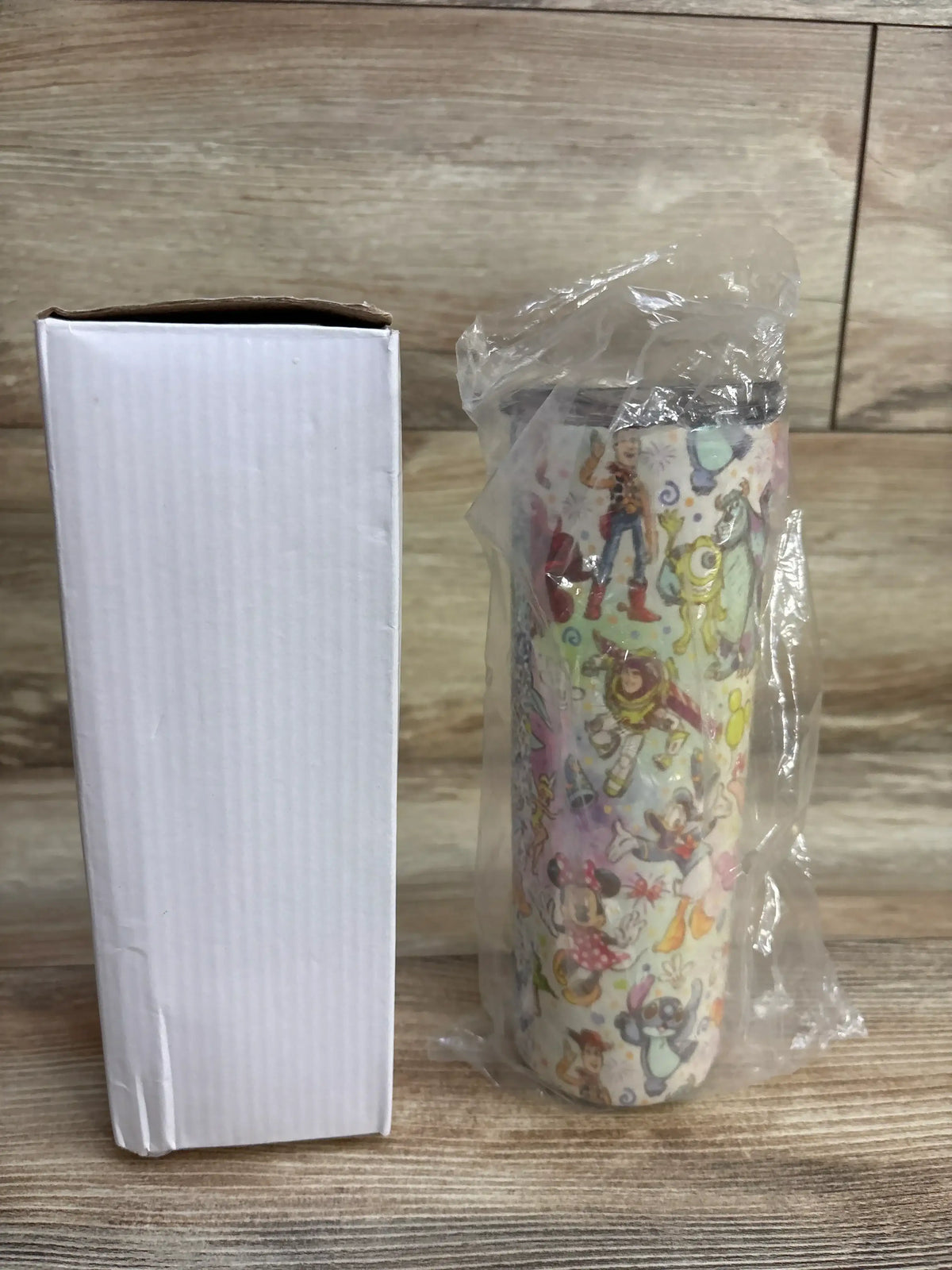 Disney-themed insulated stainless steel tumbler with cartoon characters wrapped in plastic next to plain white box on wood surface